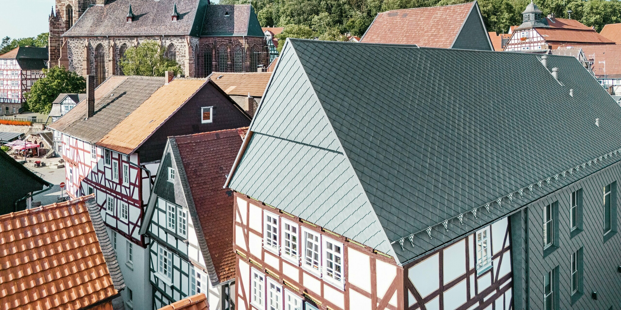 Das Dach des sanierten Volkshauses in Homberg, DE, mit PREFA Aluminiumrauten 29 × 29 in P.10 Steingrau, eingebettet in die historische Altstadt mit Fachwerkhäusern, Kirche und Burg. Die langlebige und robuste Aluminiumverkleidung verbindet modernes Design mit dem Charme der historischen Umgebung. In der Bildmitte ist der Giebel des Gebäudes zu sehen, im Hintergrund die Kirche und der Burgberg. 