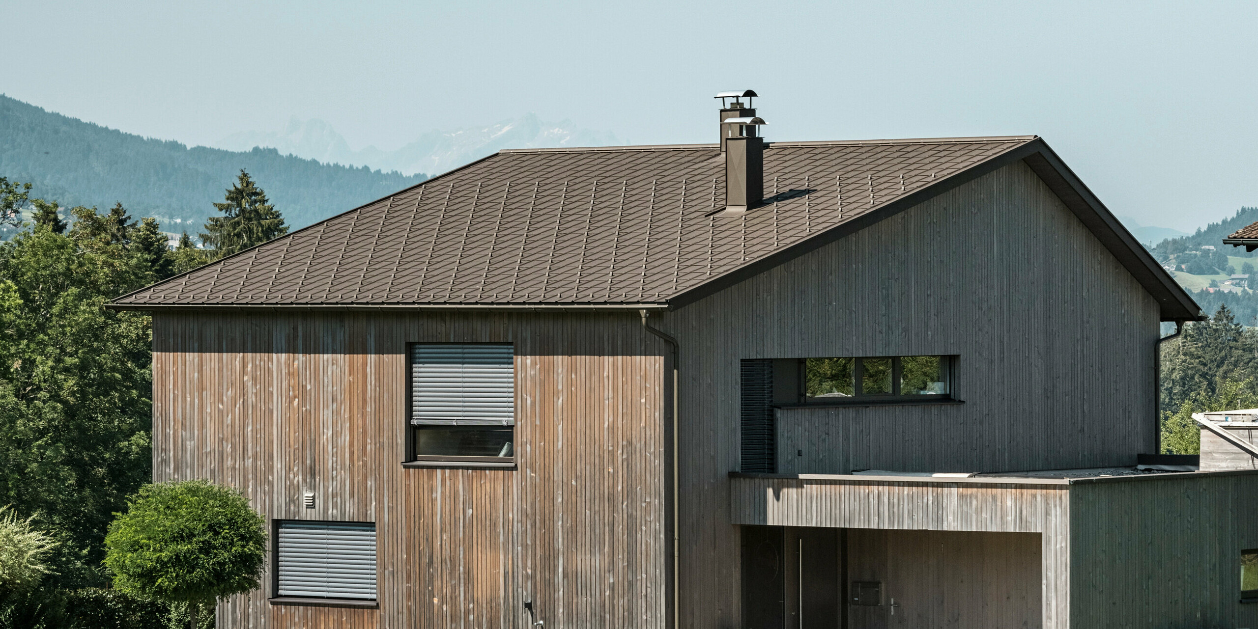 Sidebilde av en moderne enebolig i Krumbach med en kombinasjon av en naturlig trefasade og et slitesterkt PREFA-tak i aluminium i fargen P.10 brun. Det harmoniske fargevalget viser den elegante integrasjonen av moderne design og tradisjonelle materialer, og PREFAs takplater gir en estetisk og funksjonell takdesign.