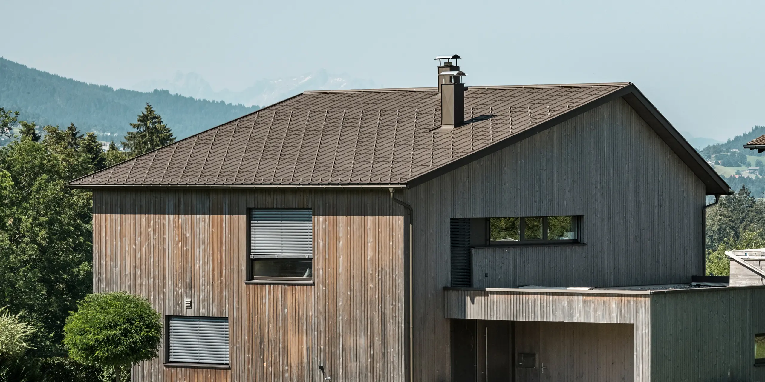 Sidebilde av en moderne enebolig i Krumbach med en kombinasjon av en naturlig trefasade og et slitesterkt PREFA-tak i aluminium i fargen P.10 brun. Det harmoniske fargevalget viser den elegante integrasjonen av moderne design og tradisjonelle materialer, og PREFAs takplater gir en estetisk og funksjonell takdesign.