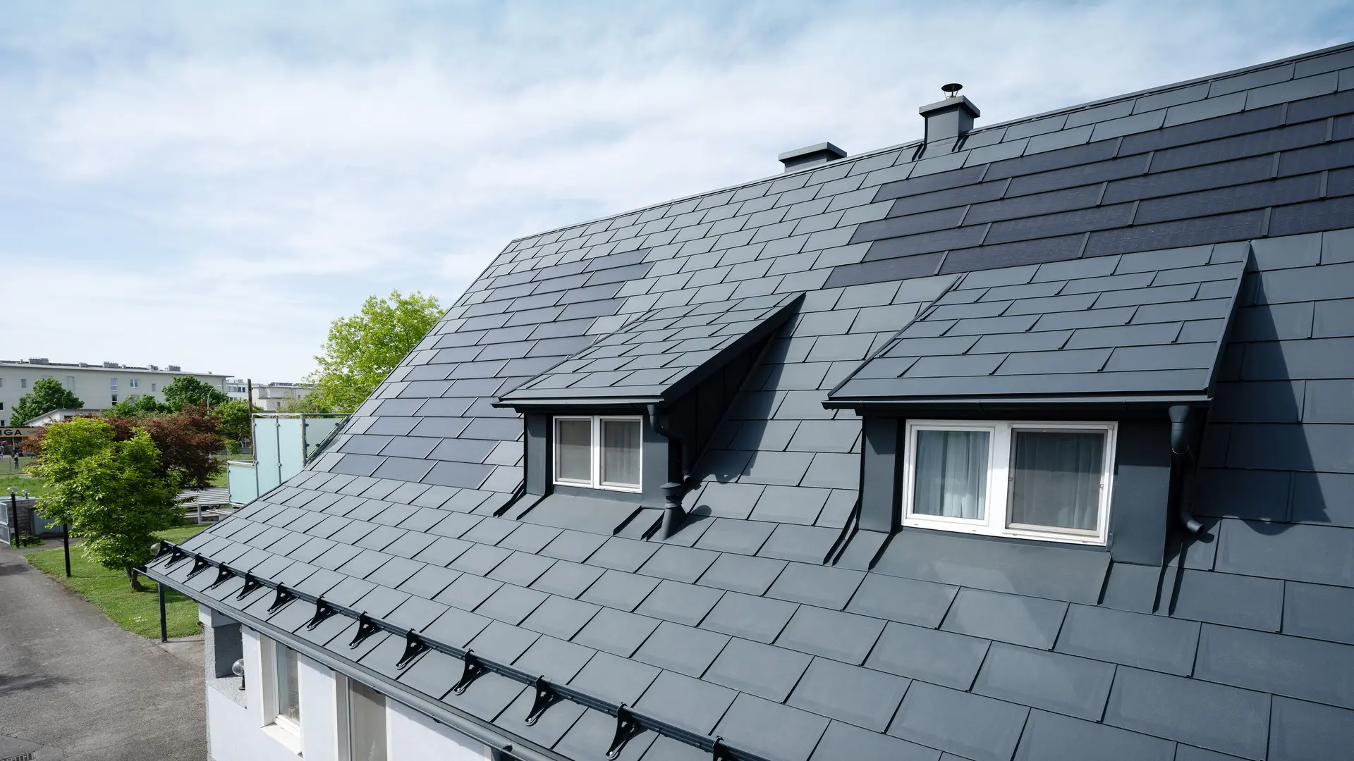 Nærbilde av et tak som er dekket med store PREFA solcelletakplater og PREFA takplate R.16 i P.10 antrasitt. To takvinduer er synlige. Solcelletakplaterne er jevnt fordelt over taket og gir en moderne, bærekraftig løsning.