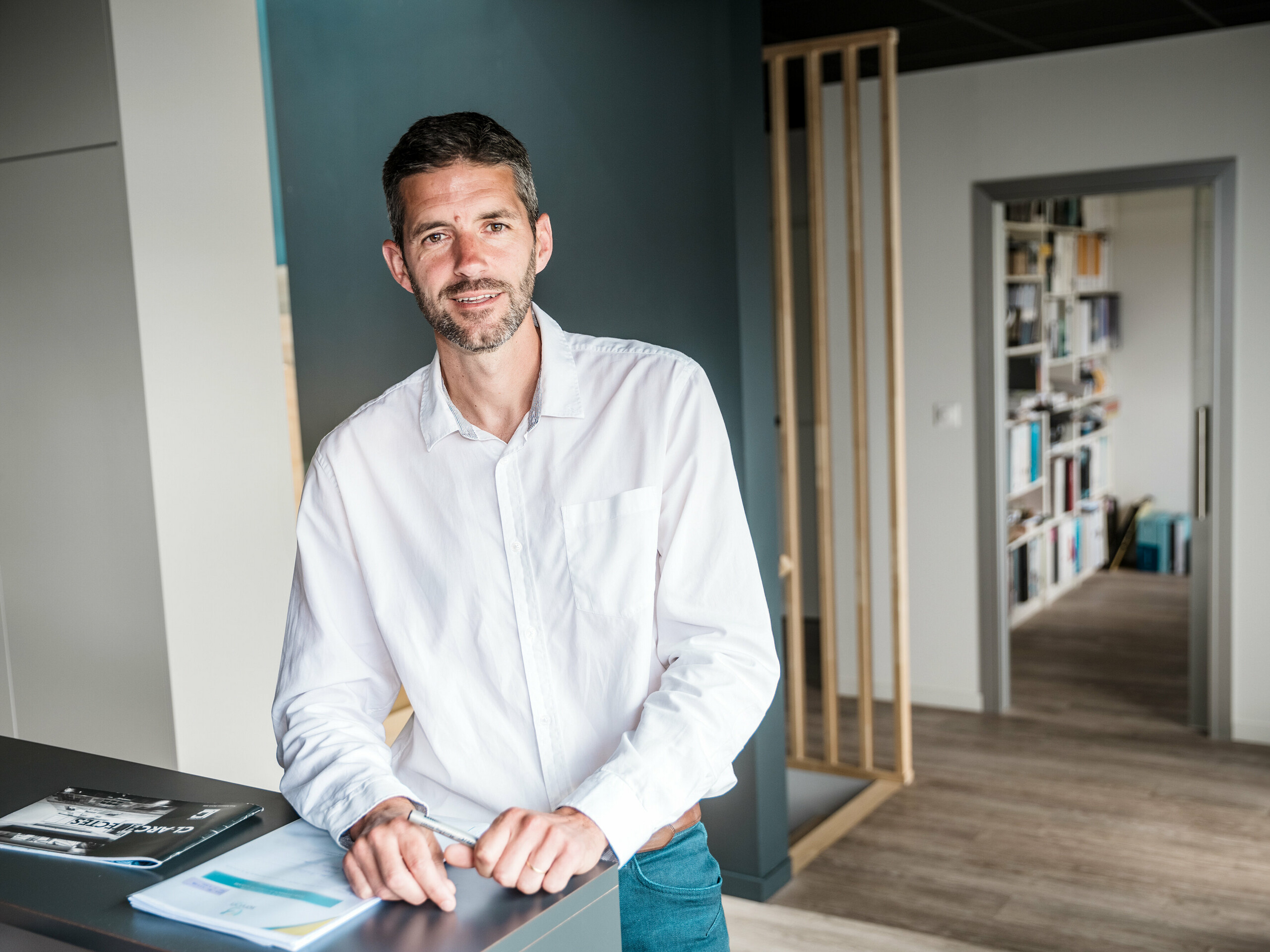 Im Vordergrund des Fotos ist der Architekt Hugues Launay vom Architekturbüro CLARC Architectes zu sehen. Er steht mittig, leicht links, und trägt formelle Kleidung, wie sie für ein Vorstellungsgespräch üblich ist. Im Hintergrund erstreckt sich die geschwungene Aluminiumfassade des Collège Léontine Dolivet, deren silbermetallische Oberfläche Lichtreflexe erzeugt. Der Mann blickt leicht zur Seite, seine Haltung wirkt präsent und selbstsicher und unterstreicht seine Rolle als Projektverantwortlicher. Die Aufnahme entstand tagsüber bei weichem, natürlichem Licht, das die Konturen deutlich hervorhebt.