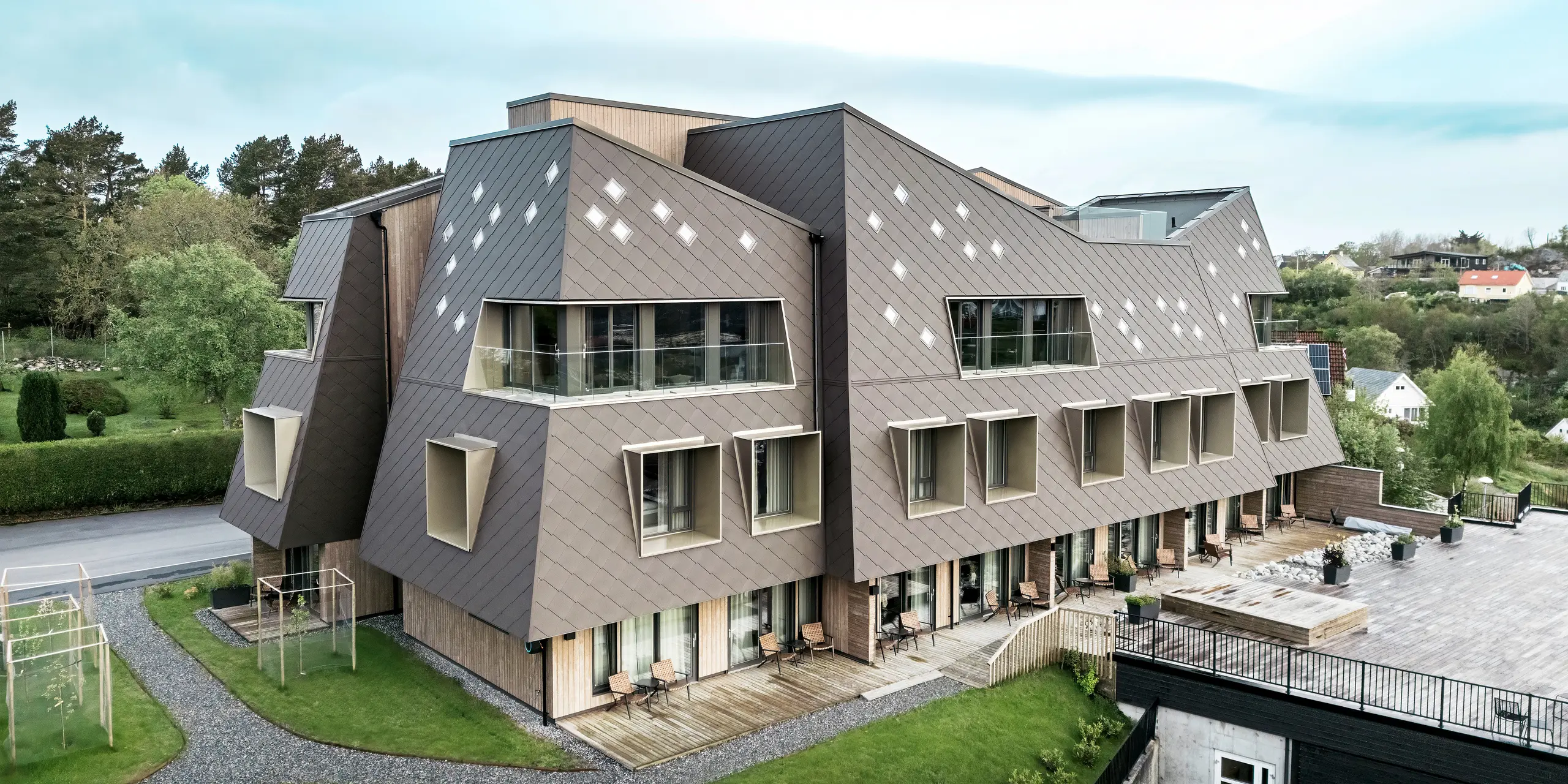 Eksteriørbilde av Beckerwyc-huset i Bekkjarvik, Norge, med fokus på de velholdte uteområdene og den markante fasaden av PREFA-veggribber 44 × 44 i valnøttbrun. Bygningens skulpturelle arkitektur, kombinert med en romslig terrasse og balkonger, skaper en harmonisk forbindelse mellom inne- og uteområdene. Den futuristiske designen og det naturlige fargevalget passer godt inn i det omkringliggende landskapet.