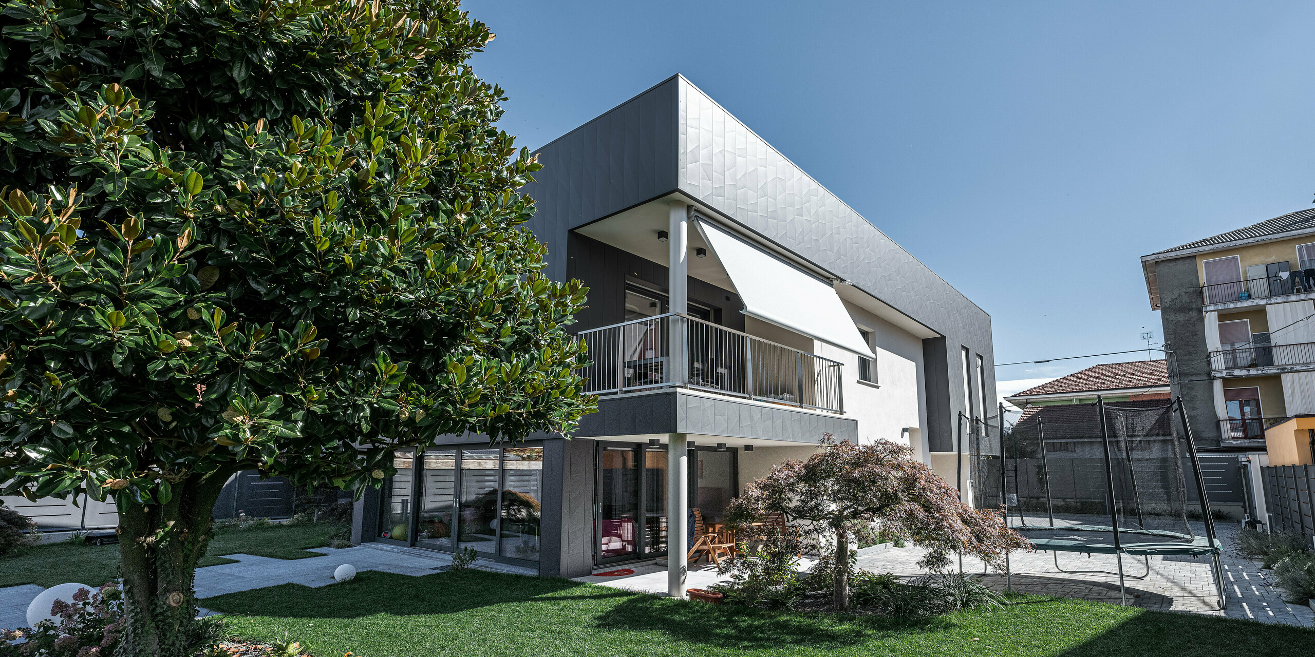 Ansicht eines modernen Einfamilienhauses in Savigliano, Italien, ausgestattet mit einer PREFA Aluminiumfassade, Modell Siding.X in P.10 Dunkelgrau. Die Fassade aus Blech verleiht dem Haus ein zeitgemäßes, geometrisch anspruchsvolles Aussehen und bietet dank der P.10 Lackierung langanhaltenden Schutz und geringen Wartungsaufwand. Das gut gepflegte Grün des Gartens und einladende Außenbereiche ergänzen die kühle Eleganz der Aluminiumfassade und schaffen einen ansprechenden Wohnraum im Freien.