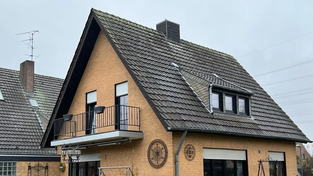 Enkeltstående hus i gul klinker med bratt sadeltak og mørk takbelegg før takrenoveringen; på hagesiden med balkong, takvindu, terrasse med sitteplasser og plen i forgrunnen.