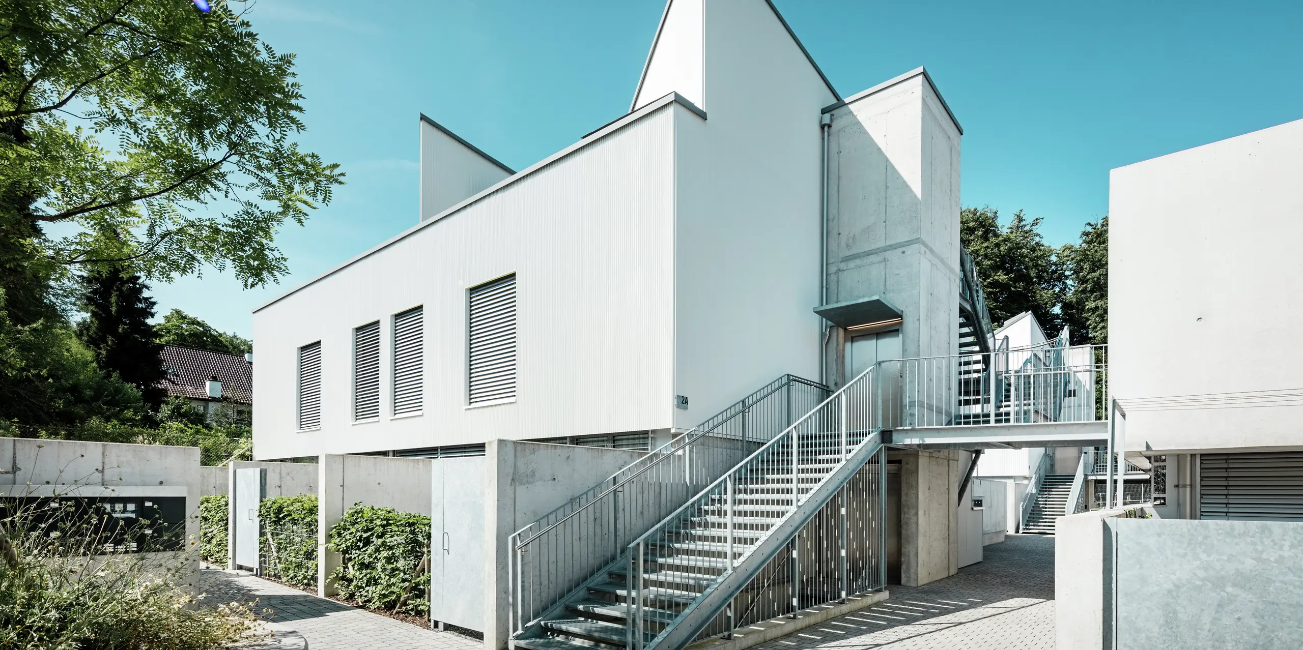 Ensemble moderner Tiny Homes mit markanten Spitzdächern, verkleidet mit dem PREFA Zackenprofil in der Farbe P.10 Reinweiß. Die schlichte, klare Fassadengestaltung harmoniert mit den Sichtbetonelementen und der umlaufenden Stahlkonstruktion. Die langlebige PREFA Aluminiumfassade schützt zuverlässig vor Witterung und sorgt für eine wartungsarme, ästhetisch reduzierte Optik.