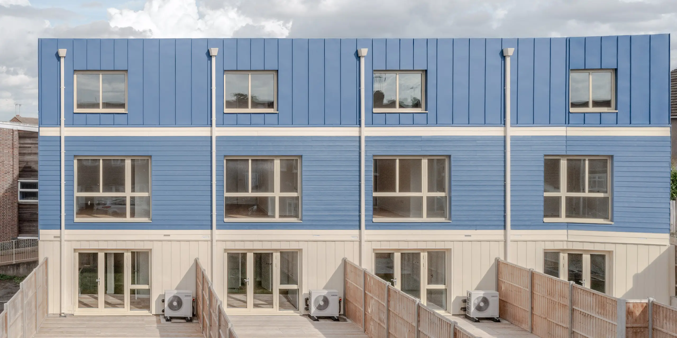 Die Gartenseite der Reihenhäuser in Goldsmith Mews zeigt die markante taubenblaue PREFA FALZONAL Fassaden- und Dachgestaltung, die den Gebäuden einen klaren, modernen Charakter verleiht. Die präzisen vertikalen Falze und die blauen Paneele erzeugen ein rhythmisches, lebendiges Fassadenbild. Die hellen Erdgeschossbereiche und großen Fenster öffnen die Architektur nach außen. Das hochwertige PREFA Aluminium bietet dauerhaften Witterungsschutz, hohe Farb- und Formstabilität und ermöglicht eine langlebige, ästhetische Gestaltung mit hoher Wiedererkennung.