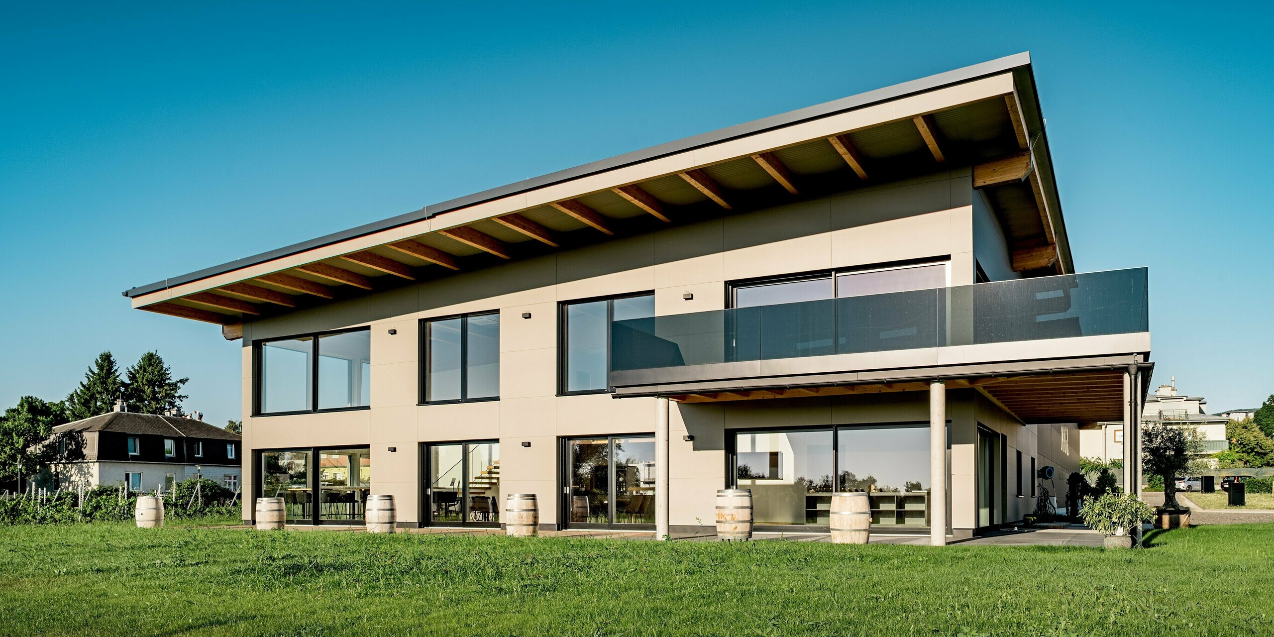 Frontalansicht des modernen Weinguts Nigl in Perchtoldsdorf mit klarer Linienführung und PREFA Aluminiumfassade in P.10 Prefabronze. Die edlen Verbundplatten strukturieren die Fassade präzise und harmonieren perfekt mit den bodentiefen Fenstern, der gläsernen Balkonbrüstung und dem weit auskragenden Pultdach mit sichtbaren Holzbalken. Auf der Terrasse sorgen dekorative Weinfässer für regionale Akzente. Die umliegenden Weingärten und das satte Grün der Wiese unterstreichen die naturnahe Lage. Die PREFA Fassade überzeugt mit langlebiger Qualität, Witterungsbeständigkeit und moderner Optik.