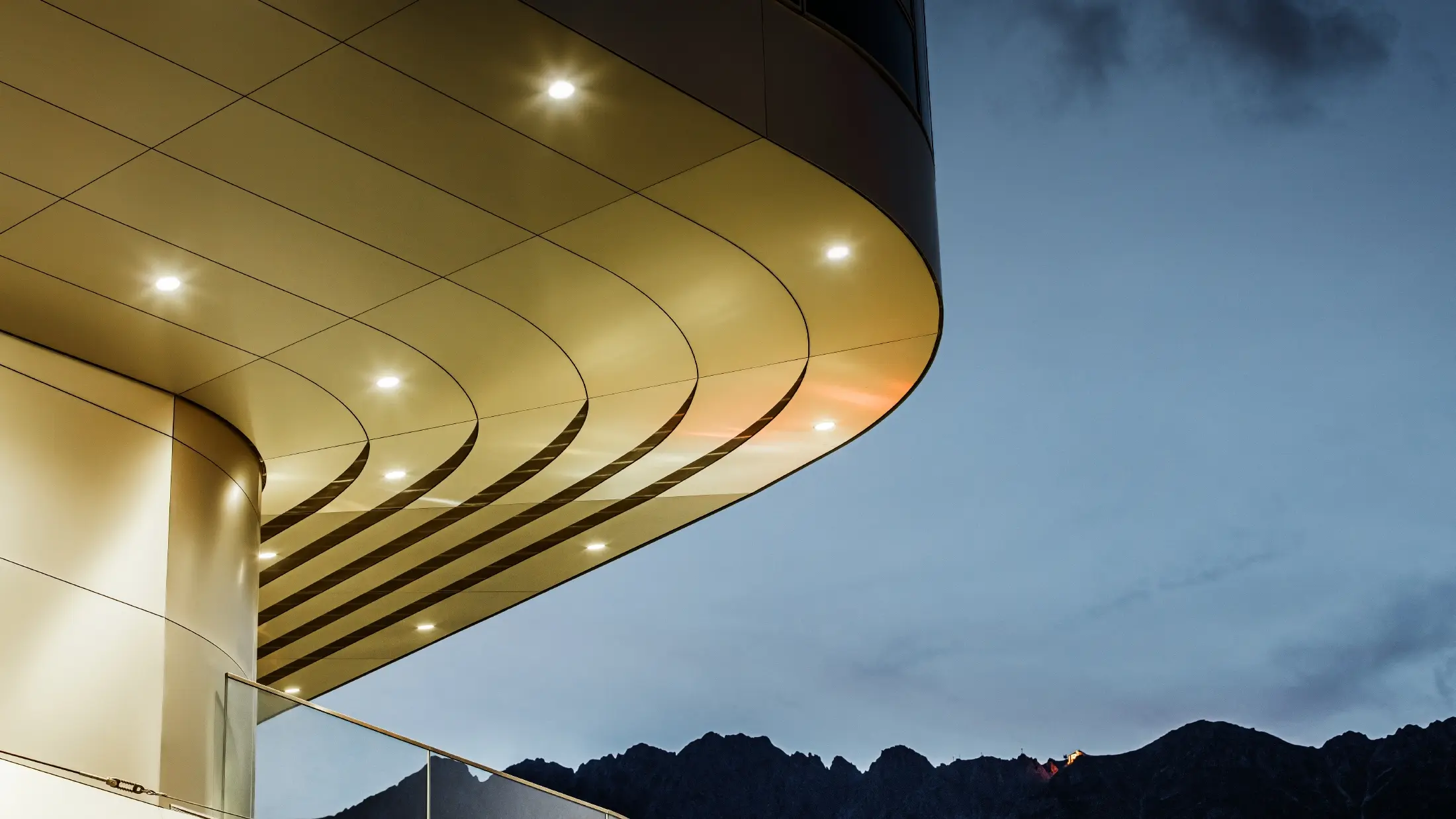 Bildet viser en imponerende arkitektonisk detaljvisning av PREFA aluminiums-komposittplater i fargen børstet aluminium. Platen er svakt buet og har elegante, flytende linjer som fremheves spesielt godt ved målrettet belysning om kvelden. Platens rene struktur og elegante aluminiumspreg kompletterer det moderne fasadedesignet og tilbyr en elegant, tidsriktig løsning for arkitektur av høy kvalitet.