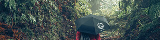Fotoet  i skogen av en kvinnelig turgåer i rød jakke med PREFA paraply og treningsbag, symboliserer PREFA miljøvern og bærekraft, samt sirkulærøkonomi og resirkulering
