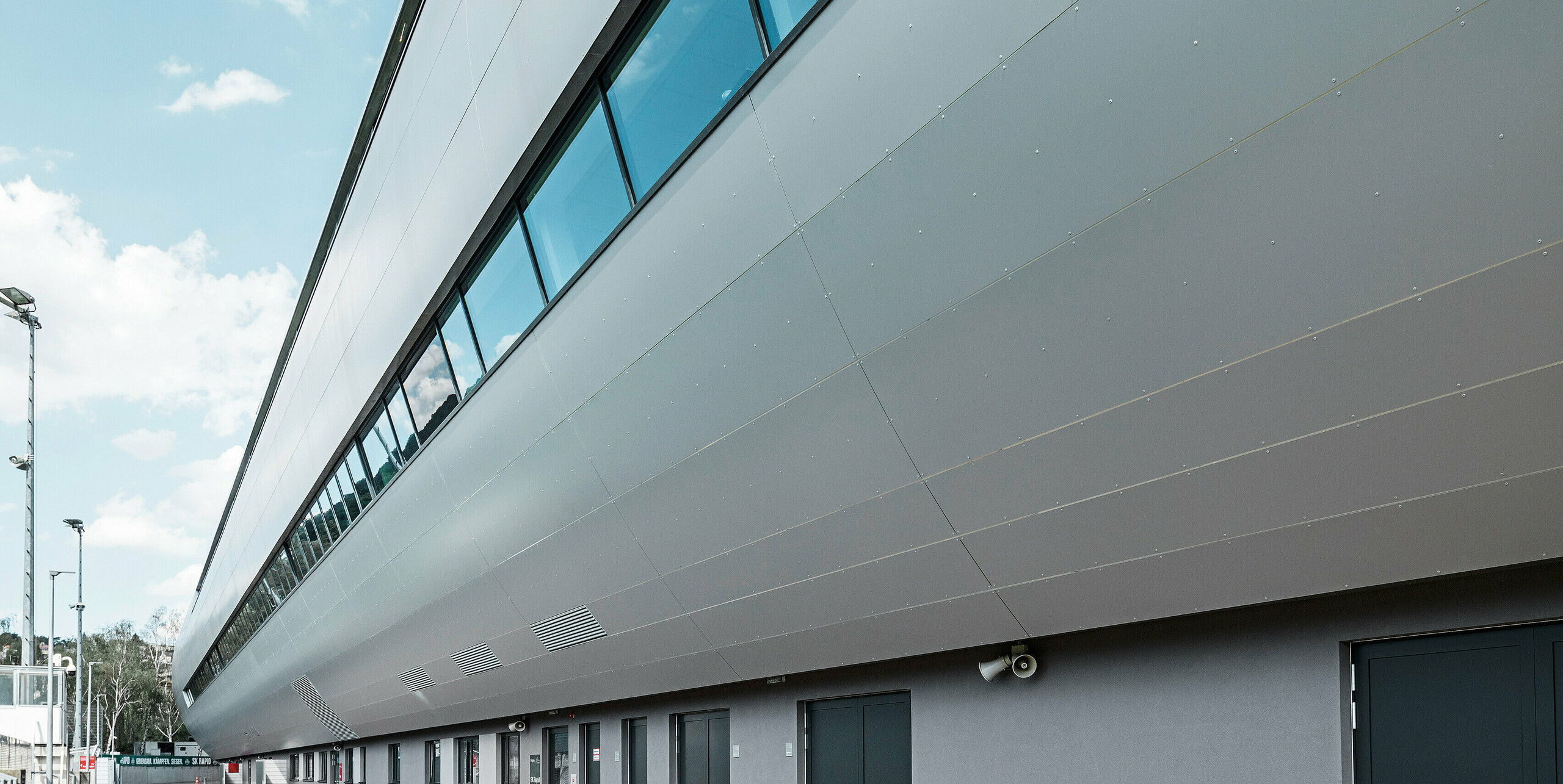 Seitenansicht des Allianz Stadions von SK Rapid Wien, verkleidet mit PREFA Aluminium Verbundplatten in Silbermetallic. Die Fassade zeigt die beeindruckende Länge und klare Linienführung des Bauwerks, während die großflächigen Fenster den Blick zum Trainingsplatz rahmen. In den Fensterscheiben reflektiert der blaue Himmel. Die hochwertige Metallverkleidung verkörpert Beständigkeit und moderne Architektur.