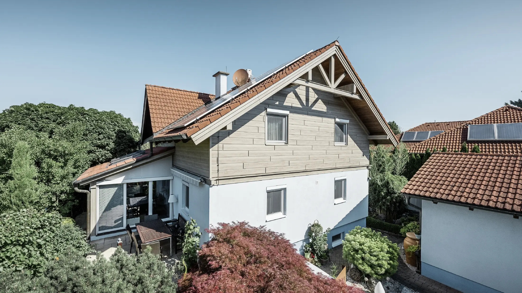 Enkeltstående hus med sadeltak og rødt takbelegg, kombinert med en moderne fasadedesign. Den øvre gavlen er kledd med grå PREFA-fasadepaneler, mens første etasje har en hvit pussfasade. På taket er det solcellepaneler. Huset ligger i en grønn hage med terrasse, hagemøbler og frodig beplantning, omgitt av andre bolighus med lignende takarkitektur.