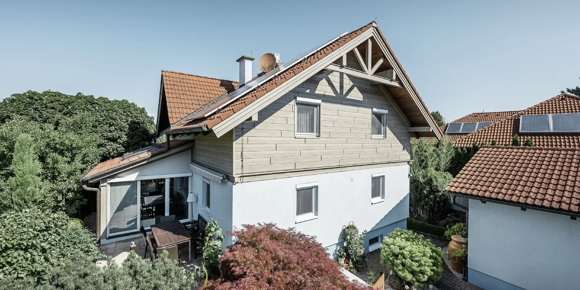 Enkeltstående hus med sadeltak og rødt takbelegg, kombinert med en moderne fasadedesign. Den øvre gavlen er kledd med grå PREFA-fasadepaneler, mens første etasje har en hvit pussfasade. På taket er det solcellepaneler. Huset ligger i en grønn hage med terrasse, hagemøbler og frodig beplantning, omgitt av andre bolighus med lignende takarkitektur.