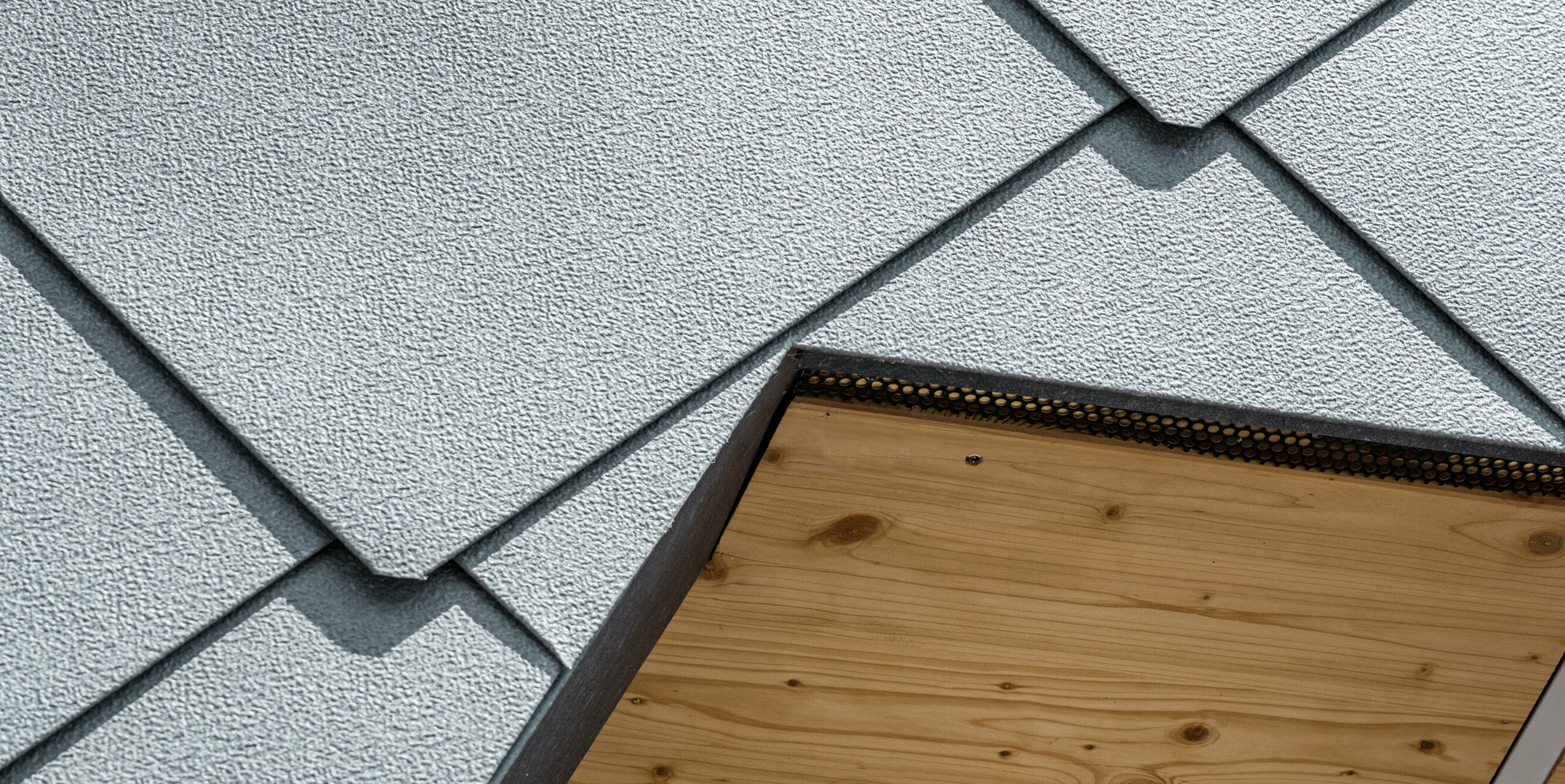 Detailaufnahme der Schnittstelle zwischen Holz und Aluminium am Sportgebäude FC Prilly-Sports in der Schweiz. Die robuste Oberfläche der PREFA Dachrauten 44x44 in P.10 Steingrau wurde durch die bewährte Stucco-Prägung verfestigt, wodurch das Aluminium unempfindlicher gegen Bearbeitungsspuren ist. Als Gegenstück dazu ist auf der Unterseite des Daches die natürliche Holzverkleidung zu erkennen. Diese Nahaufnahme zeigt die sorgfältige Handwerkskunst und Präzision, mit der die robusten, wetterbeständigen Aluminiumelemente in die architektonische Struktur der Sportanlage integriert sind. Zudem wird die ästhetische Verbindung von modernen Baustoffen und traditionellen Materialien betont.