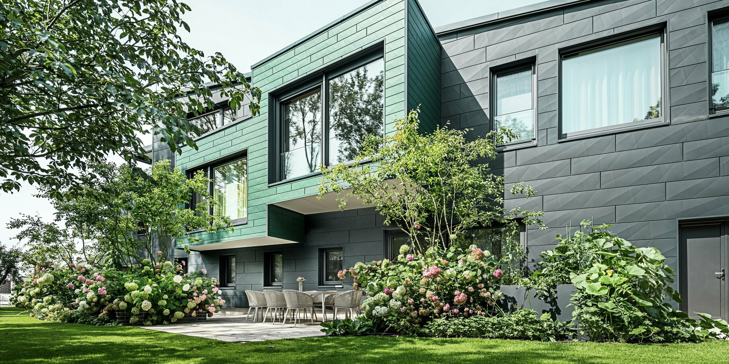 Das Einfamilienhaus in Türkheim zeigt eine harmonische Kombination aus PREFA Siding X-Paneelen in den Farben Anthrazit und Moosgrün. Die markanten Fassadenelemente mit ihren markanten Kanten schaffen eine lebendige Optik und fügen sich perfekt in die grüne Umgebung ein. Im Vordergrund ist ein stilvoller Gartenbereich mit einer Sitzgruppe aus modernen Stühlen und üppigem Blumenschmuck zu sehen. Die Pflanzen, insbesondere die rosa Hortensien, setzen einen schönen Kontrast zur dunklen Fassade. Die langlebige und pflegeleichte Aluminiumfassade schützt das Gebäude zuverlässig vor Witterungseinflüssen und sorgt für eine zeitlose Ästhetik.