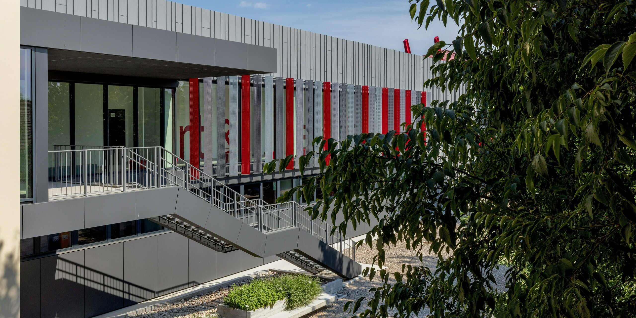 Die lebendige Außenanlage der Scuola Arte e Mestieri F. Bertazzoni in Suzzara besticht durch eine Außentreppe, die an der PREFA-Aluminiumfassade in Dunkelgrau und Patinagrau entlang zur Eingangsebene führt. Rhythmisch gesetzte rote Fassadenstreifen und perforierte Elemente verleihen dem Gebäude eine pulsierende Energie. In den Innenräumen unterstreichen Großbuchstaben in Rot den unverwechselbaren Charakter des Schulgebäudes. Die PREFA Produkte PREFALZ und PREFABOND überzeugen mit makellos verarbeiteten Oberflächen, höchster Formstabilität und dauerhafter Witterungsbeständigkeit. Sie sind in eine einladende Grünanlage mit mediterranem Charme eingebettet.