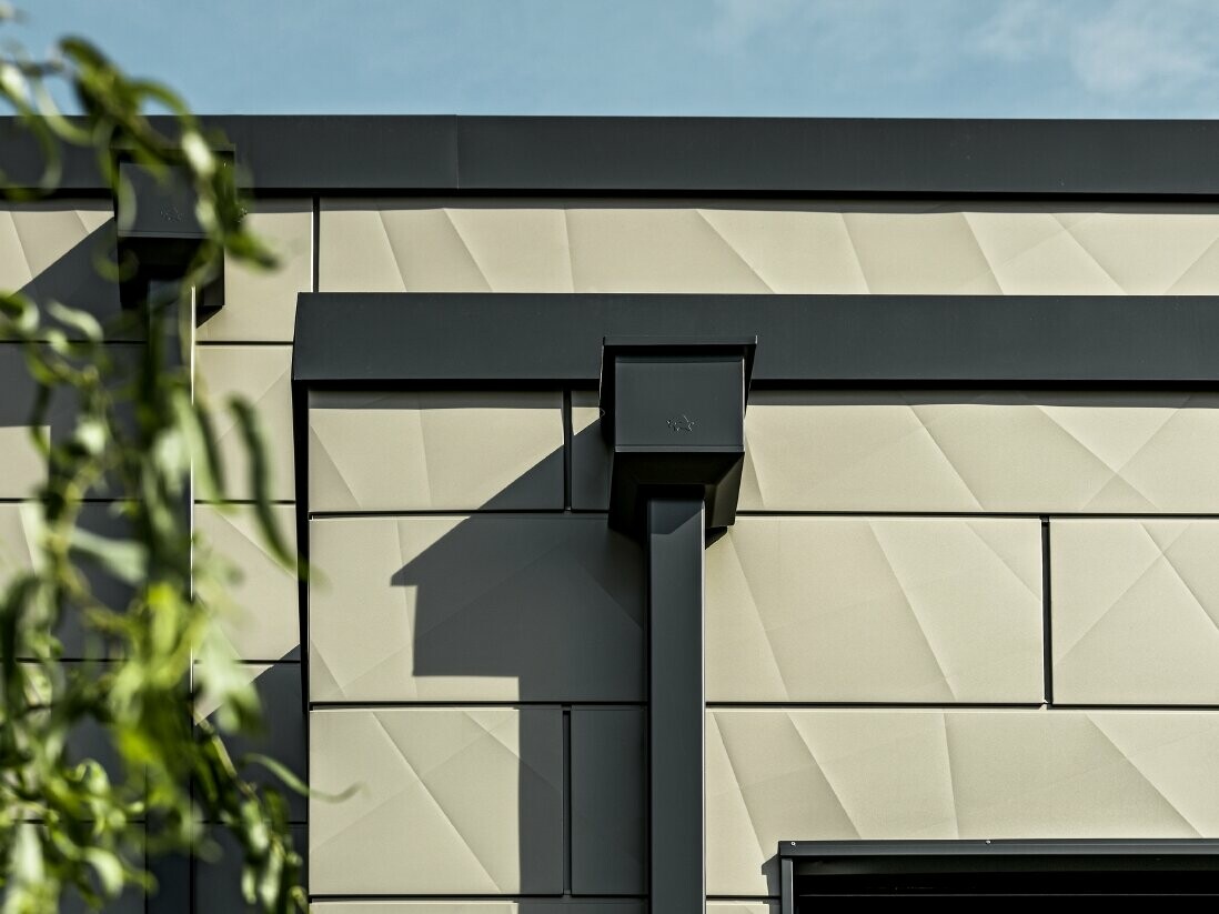 Moderne enebolig med flatt tak og smale vinduer. Fasaden er kledd med PREFA Siding.X-fasade med foldet utseende i fargen bronse. Forbindelsene ble laget i antrasitt. PREFA firkantrør med tilhørende vannoppsamlingsbeholder i antrasitt brukes til takdrenering.