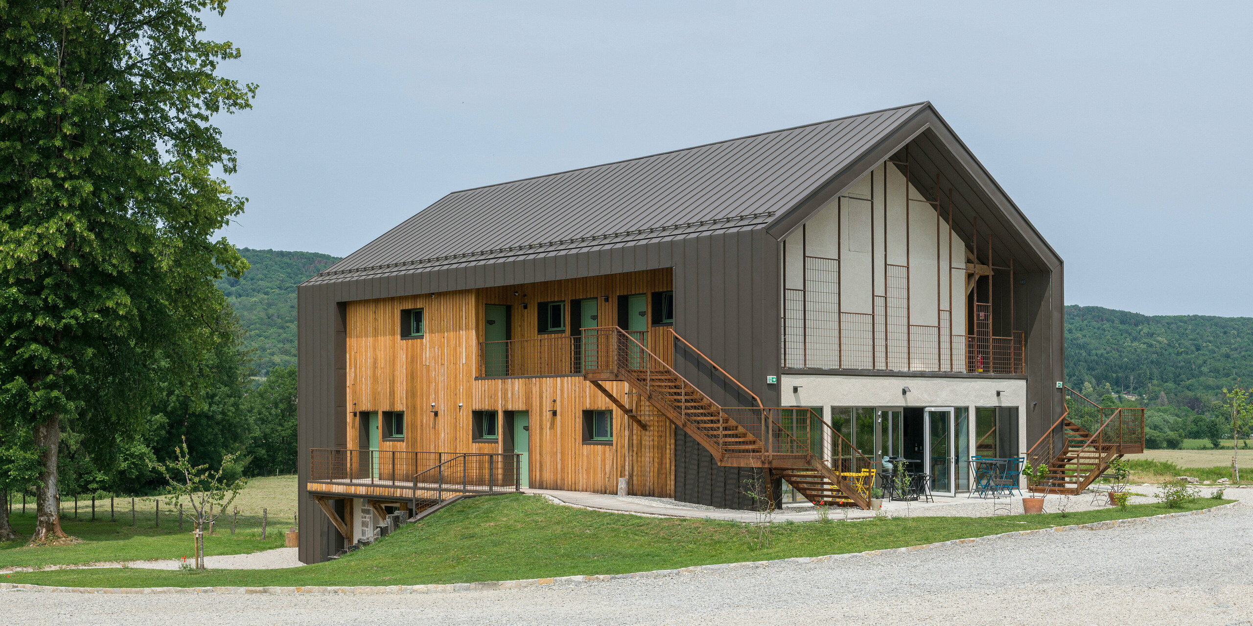 Eckansicht eines zeitgenössischen zweigeschossigen Hotels mit einer Hülle aus PREFA Aluminium und naturbelassener Holzfassade. Das Dach sowie Teile der Fassade wurden mit PREFALZ in P.10 Braun verkleidet. Hervorstechende Merkmale des Gebäudes sind große Fensterflächen sowie eine außenliegende Treppe, die zum oberen Stockwerk führt. Zudem verfügt das Hôtel en Tilleul über eine Terrasse im Erdgeschoss. Das Gebäude steht in einer idyllischen ländlichen Umgebung mit Blick auf sanfte Hügel. Am unteren Bildrand ist die Zufahrtsstraße aus Kies zu sehen. 