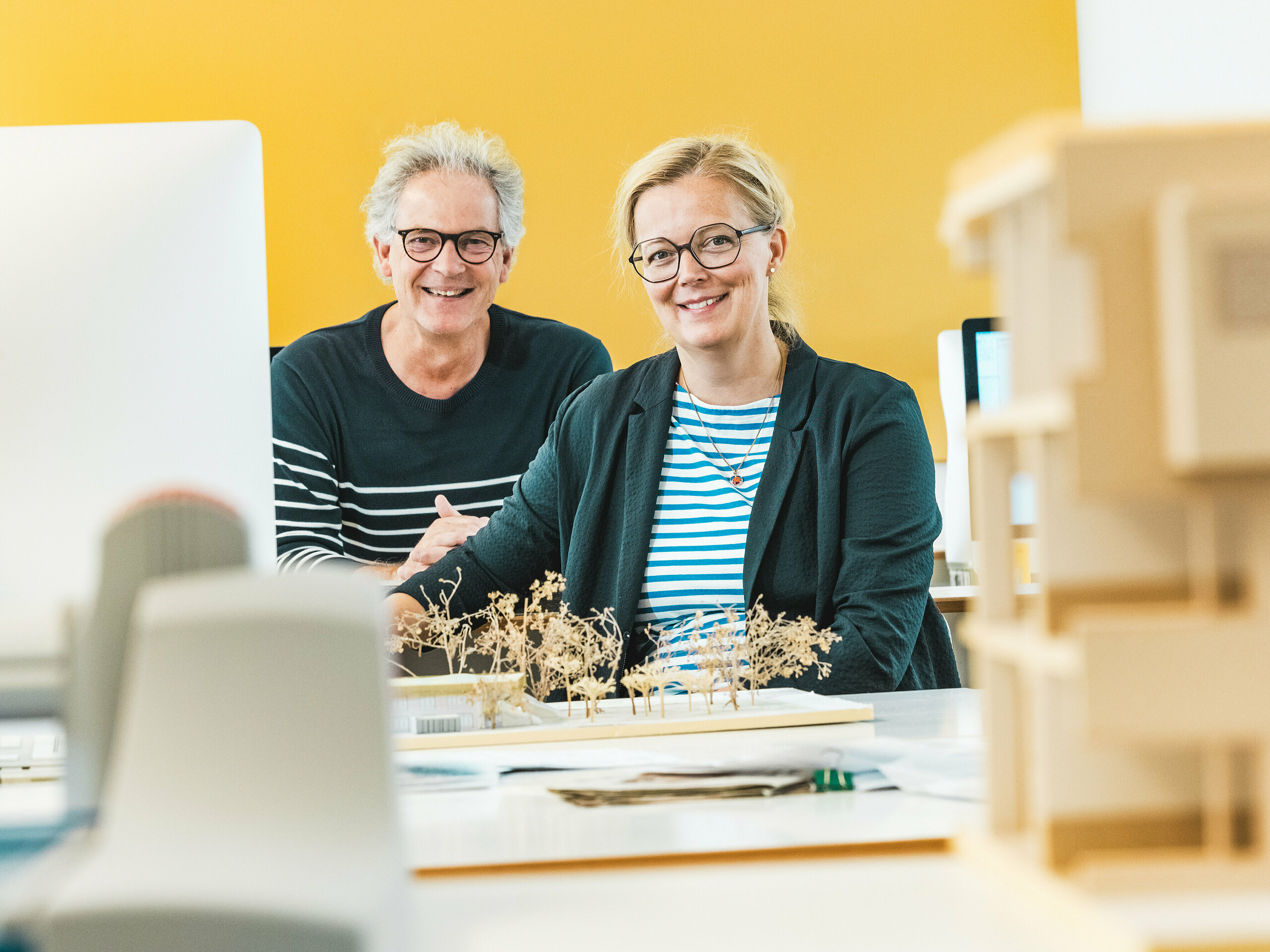 Die Architekten Claudia und Theo Härtner sitzen strahlend vor einem Architekturmodell. Das Architektenpaar realisiert die Kindertagesstätte in Alzenau, ein zukunftsweisendes Hybridgebäude mit einer Fassade aus PREFA Aluminium. Im Vordergrund des Architekturmodells sind Baumminiaturen zu sehen, die das nachhaltige Gesamtkonzept widerspiegeln. Die Kita in Alzenau kombiniert innovative Architektur mit verantwortungsvoller Stadtentwicklung und zeigt die Vielseitigkeit von PREFA Wandrauten 44 × 44 in Bronze. Die langlebige, wartungsarme und zu 100 % recycelbare Aluminiumfassade verbindet den Wohn- und den Kitabereich optisch zu einer harmonischen Einheit. Das Bild fängt die kreative Energie des Planungsprozesses ein und unterstreicht die ästhetischen und ökologischen Vorteile von PREFA-Produkten.
