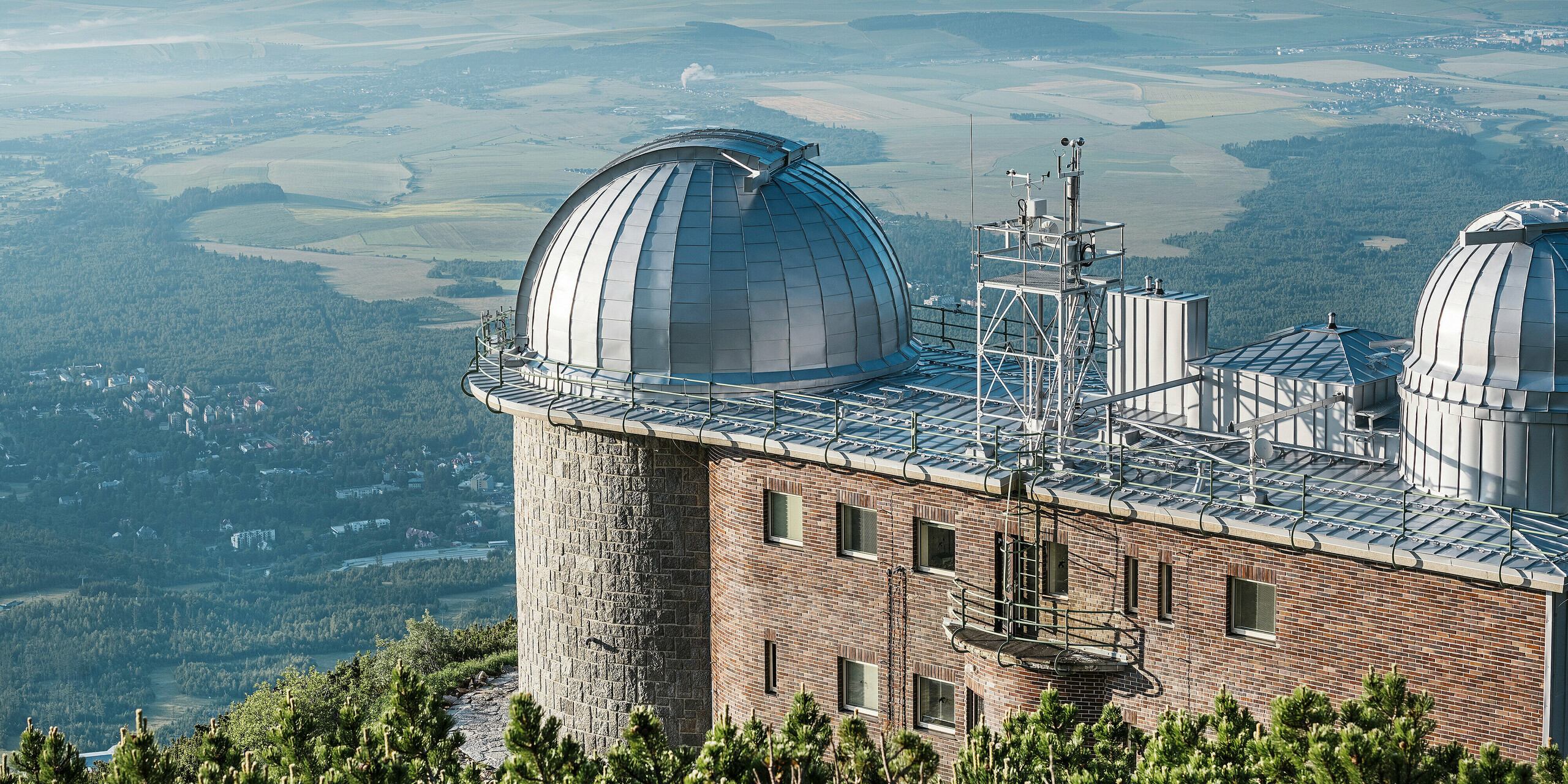 Observatorium Skalnaté Pleso, über der idyllischen slowakischen Landschaft gelegen, mit glänzenden PREFALZ Kuppeln in Silbermetallic. Das imposante Gebäude fügt sich nahtlos in seine natürliche Umgebung ein und thront majestätisch auf einem Bergkamm. Dank der langlebigen PREFA Aluminiumverkleidung, die auch extremen Witterungsbedingungen standhält, bleibt die historische Sternwarte Tag und Nacht ein Hüter der Sterne. Das robuste und langlebige Material PREFA unterstreicht die Verpflichtung zu Qualität und Nachhaltigkeit, während die Architektur eine Brücke zwischen Himmel und Erde schlägt.