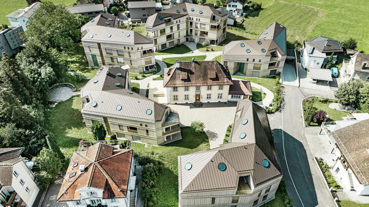 Luftaufnahme der modernen Wohnanlage in Liebwylen, Schweiz. Die Gebäude sind mit PREFALZ Aluminiumdächern in Braun gestaltet und verfügen über runde Dachfenster. Im Zentrum steht die historische Villa, umgeben von harmonisch angeordneten Neubauten, eingebettet in eine grüne Landschaft.