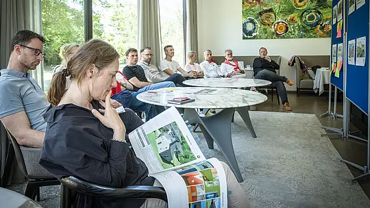 PREFARENZEN 2027 Expertendiskussion in einem hellen Seminarraum: Paulina Frey (Marketingleiterin PREFA CH) hält ein großes Architekturbuch mit Projekten aus PREFA Aluminium in Händen, während die anderen Jury-Mitglieder aufmerksam zuhören. Große Fenster lassen Tageslicht herein und bieten Blick ins grüne Freie. Die Szene vermittelt konzentrierte Analyse, kreative Diskussion und Bewertung von Design und Funktion. PREFA Aluminiumprodukte stehen dabei symbolisch für langlebige, ästhetische Dächer und Fassaden, die Innovation, Qualität und Inspiration in Architekturprojekten fördern.