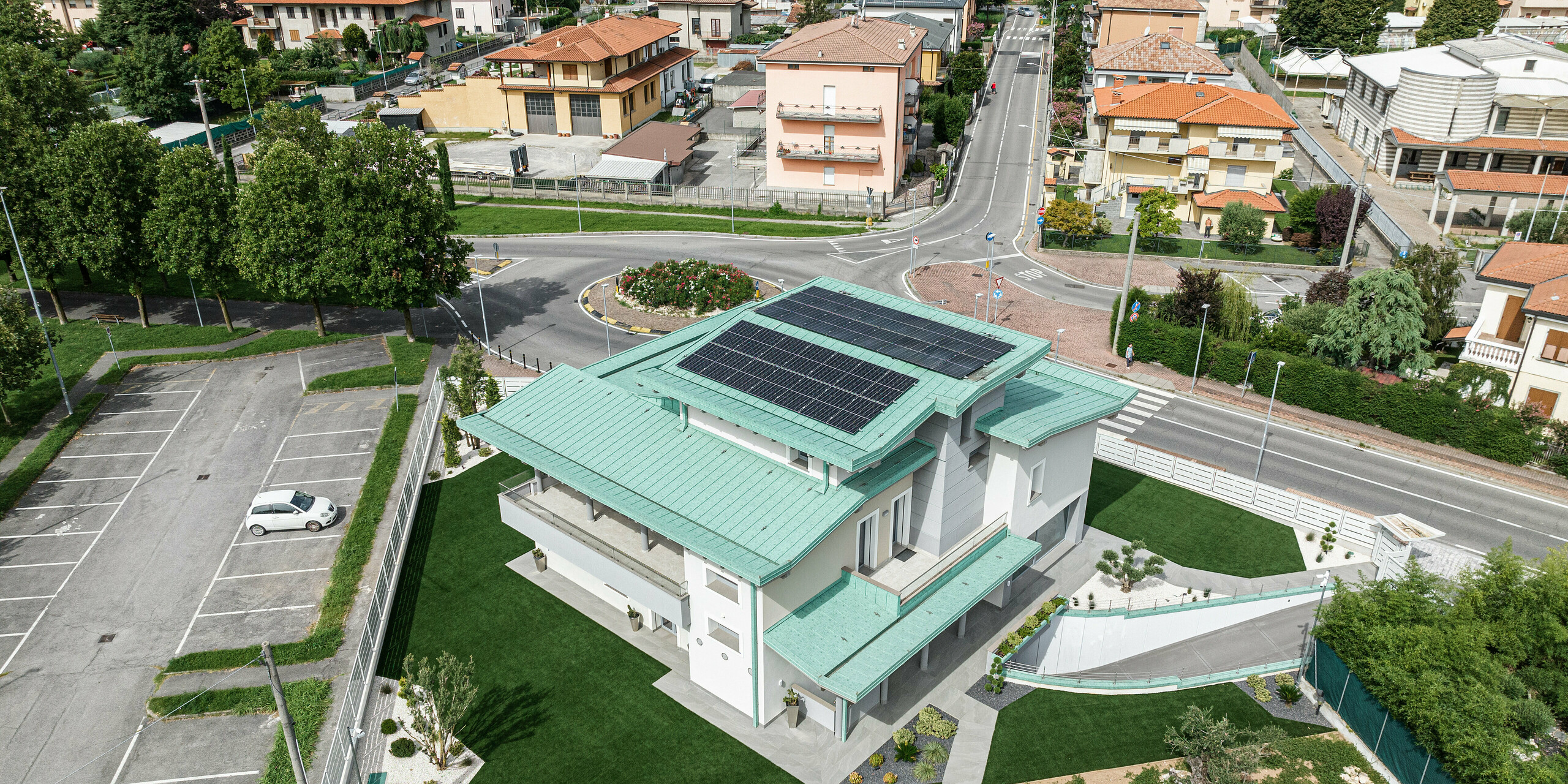 Drohnenaufnahme eines attraktiven Einfamilienhauses in Bergamo, Italien, ausgestattet mit einem nachhaltigen PREFALZ Dachsystem in der Farbe P.10 Patinagrün, ergänzt durch eine Solaranlage für Energieeffizienz. Das Gebäude steht im Kontrast zu den traditionellen roten Ziegeldächern der umliegenden Nachbarschaft und setzt ein modernes, umweltbewusstes Statement. Vor dem Gebäude befindet sich ein Kreisverkehr, dessen Abzweigungen in unterschiedliche Richtungen müden. Hinter dem Gebäude befindet sich ein großer Parkplatz, auf dem ein einziges Auto geparkt ist. Rundherum sind etliche große Bäume zu erkennen, die insbesondere in den warmen Sommermonate Schatten spenden. 