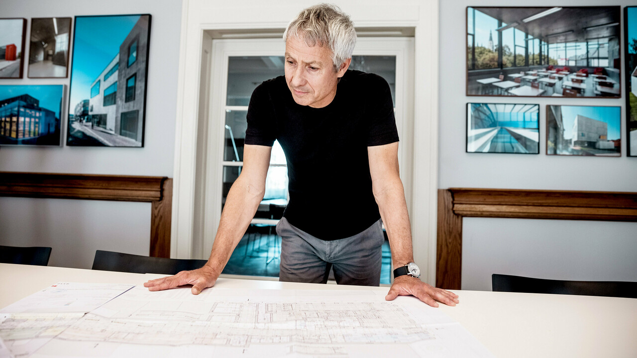 Arkitekten arbeider med detaljerte byggeplaner i et moderne kontor, omgitt av imponerende bilder av PREFA-arkitekturprosjekter.