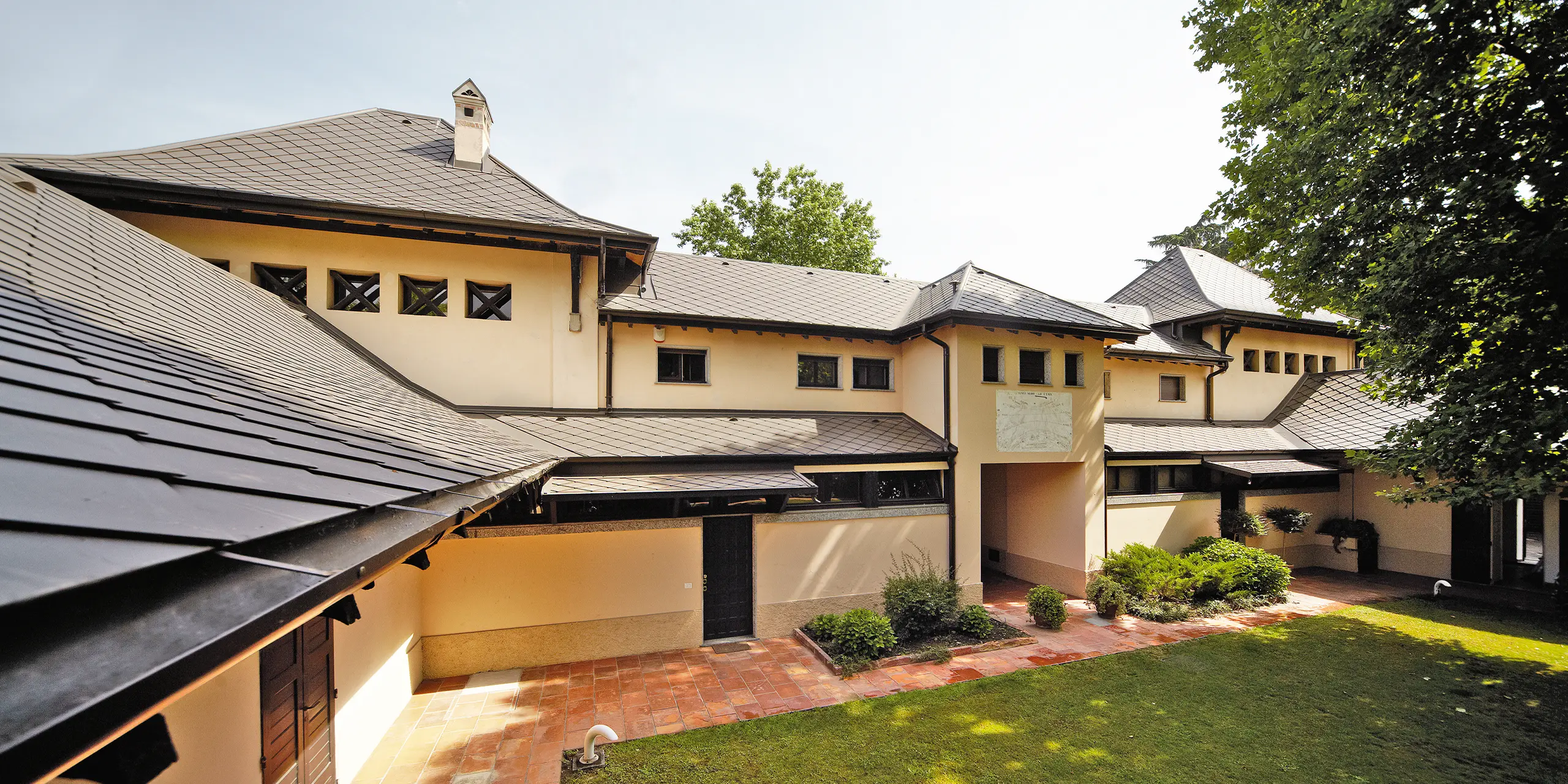 Italisensk villa med rombetak i P.10 brun fra PREFA med beige fasadefarge og bakgård med skygge