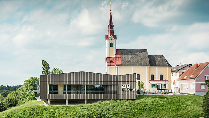 Das Veranstaltungszentrum ZIB Nikolai in St. Veit in der Südsteiermark mit einer modernen, dynamischen Aluminiumfassade in braun-beigegrauen Tönen, im Vordergrund eine grüne Wiese, im Hintergrund die Kirche von St. Nikolai mit gelber Fassade und rotem Dach.