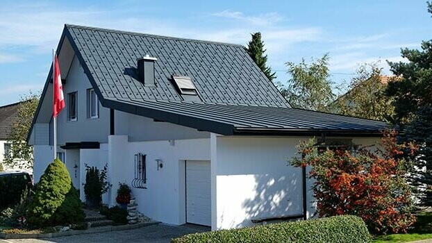 Renovert hus med saltak og tilknyttet garasje. Taket er dekket med PREFA takplate og garasjen med Prefalz i antrasitt. Foran huset er det en flaggstang med det sveitsiske flagget.