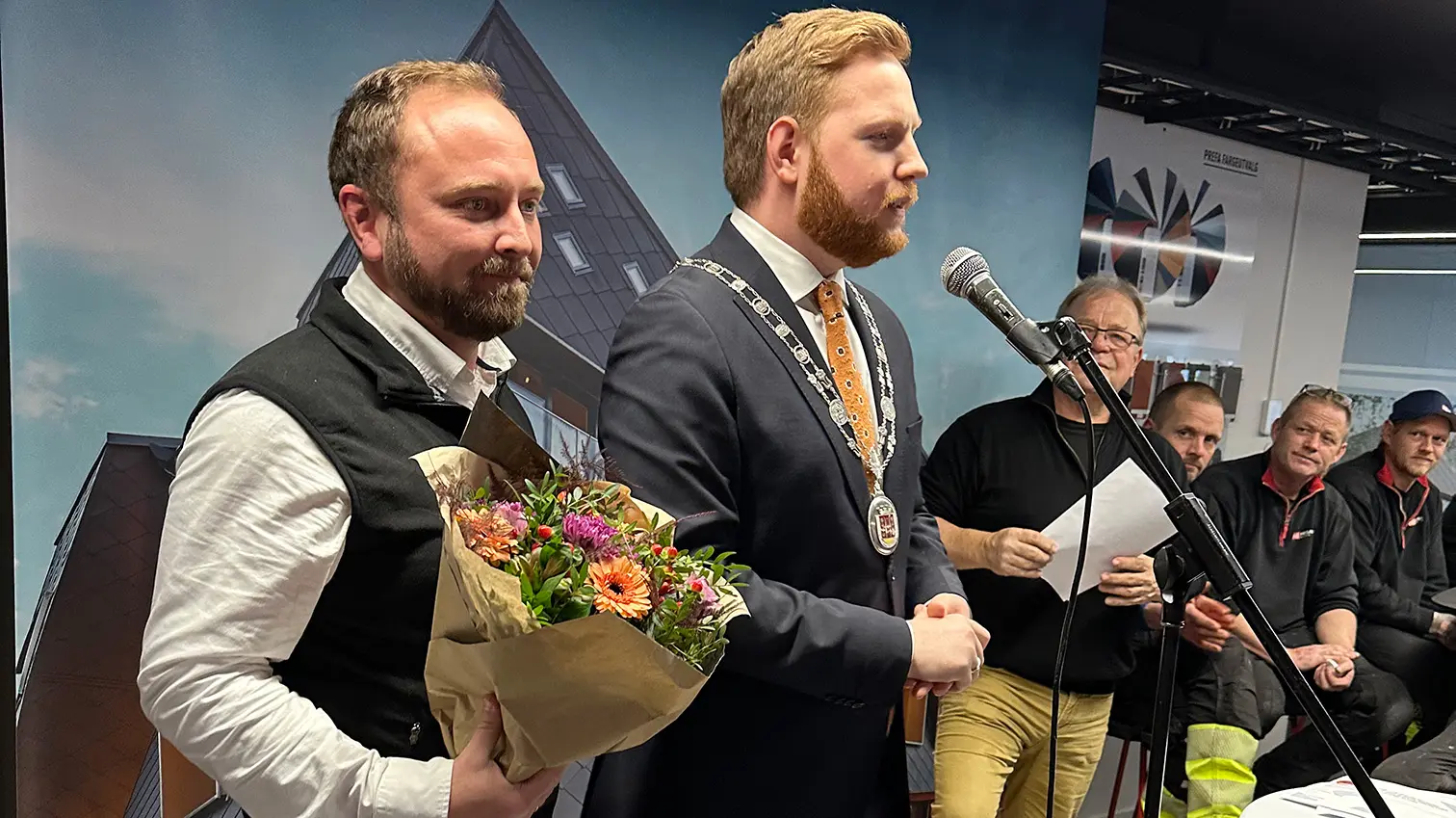 Der Bürgermeister von Skien (rechts) hält eine Rede während der Eröffnung der PREFA Academy in Skien. Neben ihm steht ein PREFA-Vertreter, der einen Blumenstrauß hält. Im Hintergrund hören weitere Teilnehmer der Veranstaltung aufmerksam zu.