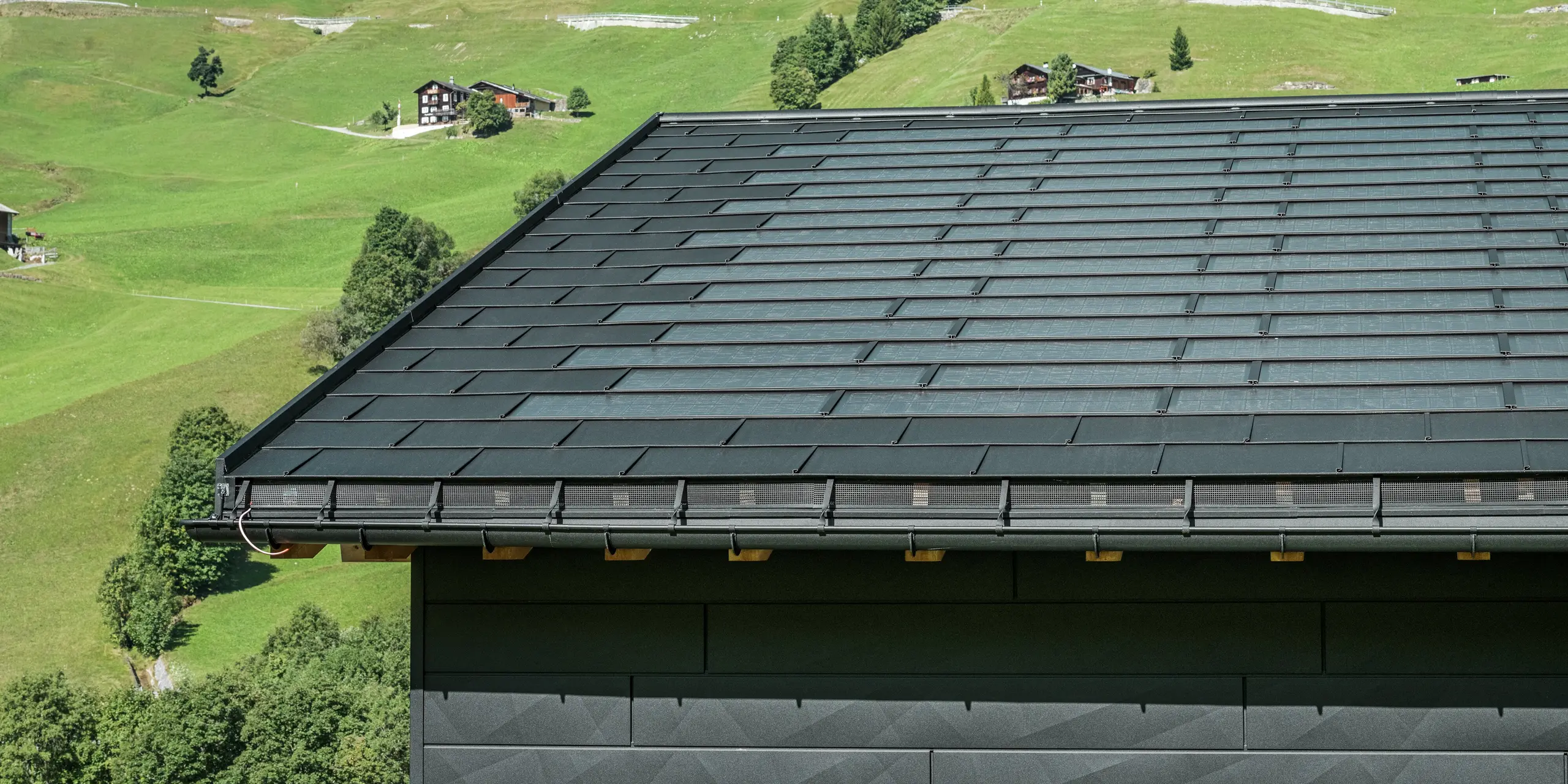 Nærbilde av takskjegget på en moderne enebolig i de alpine omgivelsene i Ascharina i Sveits. Taket er dekket med den innovative PREFA solcelle takplate og den tradisjonelle takplate R.16, begge i P.10 svart. PREFA takrenne og PREFA nedløpsrør sørger for pålitelig takdrenering. Nederst på bildet ser du de karakteristiske PREFA Siding.X fasadepaneler. De klare linjene i aluminiumsproduktene understreker det moderne designet midt i et pittoresk fjellandskap.