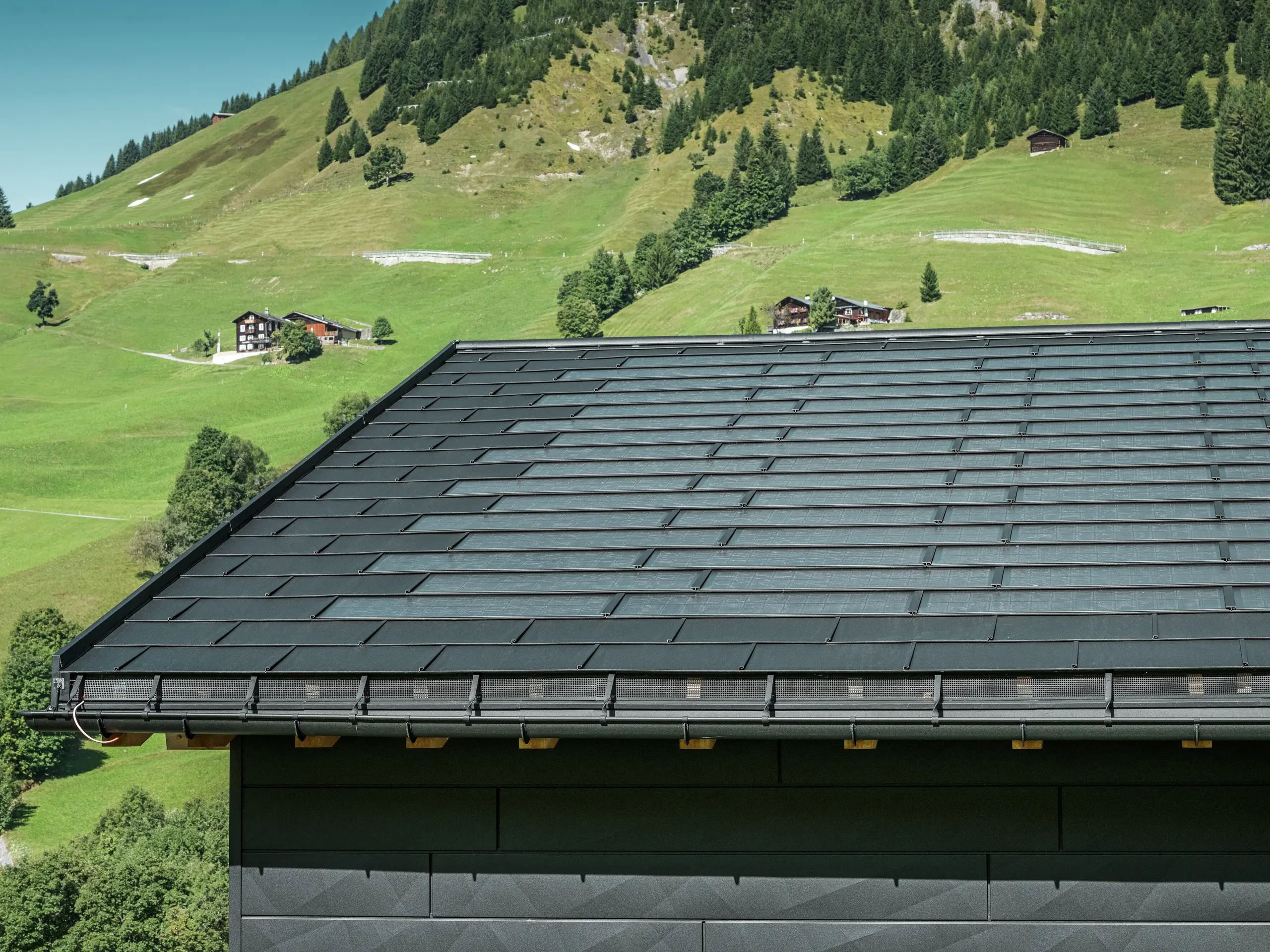 Nærbilde av takskjegget på en moderne enebolig i de alpine omgivelsene i Ascharina i Sveits. Taket er dekket med den innovative PREFA solcelle takplate og den tradisjonelle takplate R.16, begge i P.10 svart. PREFA takrenne og PREFA nedløpsrør sørger for pålitelig takdrenering. Nederst på bildet ser du de karakteristiske PREFA Siding.X fasadepaneler. De klare linjene i aluminiumsproduktene understreker det moderne designet midt i et pittoresk fjellandskap.