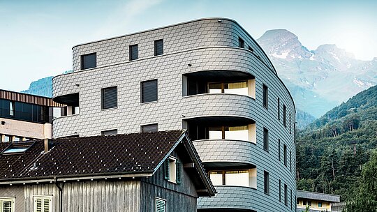Modernes, dreieckiges Wohn- und Geschäftshaus in Buchs mit markanter, abgerundeter Architektur und schuppenartiger Fassade aus PREFA Wandschindeln in P.10 Hellgrau. Die gleichmäßig umlaufende Aluminiumfassade erzeugt einen lebendigen Reptilien-Look und setzt sich als eleganter Eyecatcher vor alpiner Bergkulisse in Szene. Dank des leichten, witterungsbeständigen und langlebigen PREFA Aluminiums sind selbst enge Radien und Rundungen präzise umgesetzt – für eine homogene, wartungsarme Gebäudehülle mit zeitlos moderner Ausstrahlung.