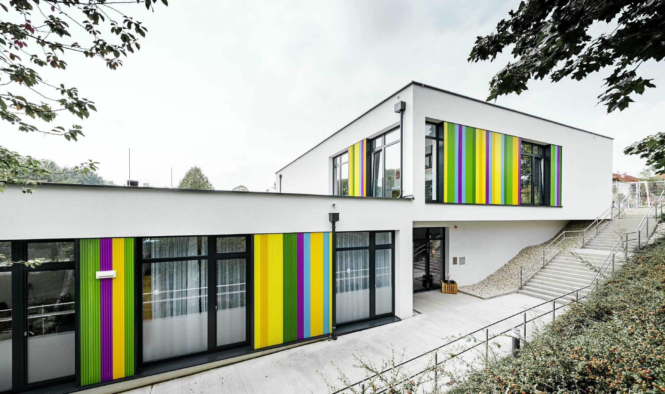 Fasadeelementer i flere farger fra PREFA på barnehagen i Hargelsberg. Bygningen har flatt tak og store vinduer helt ned til bakken.