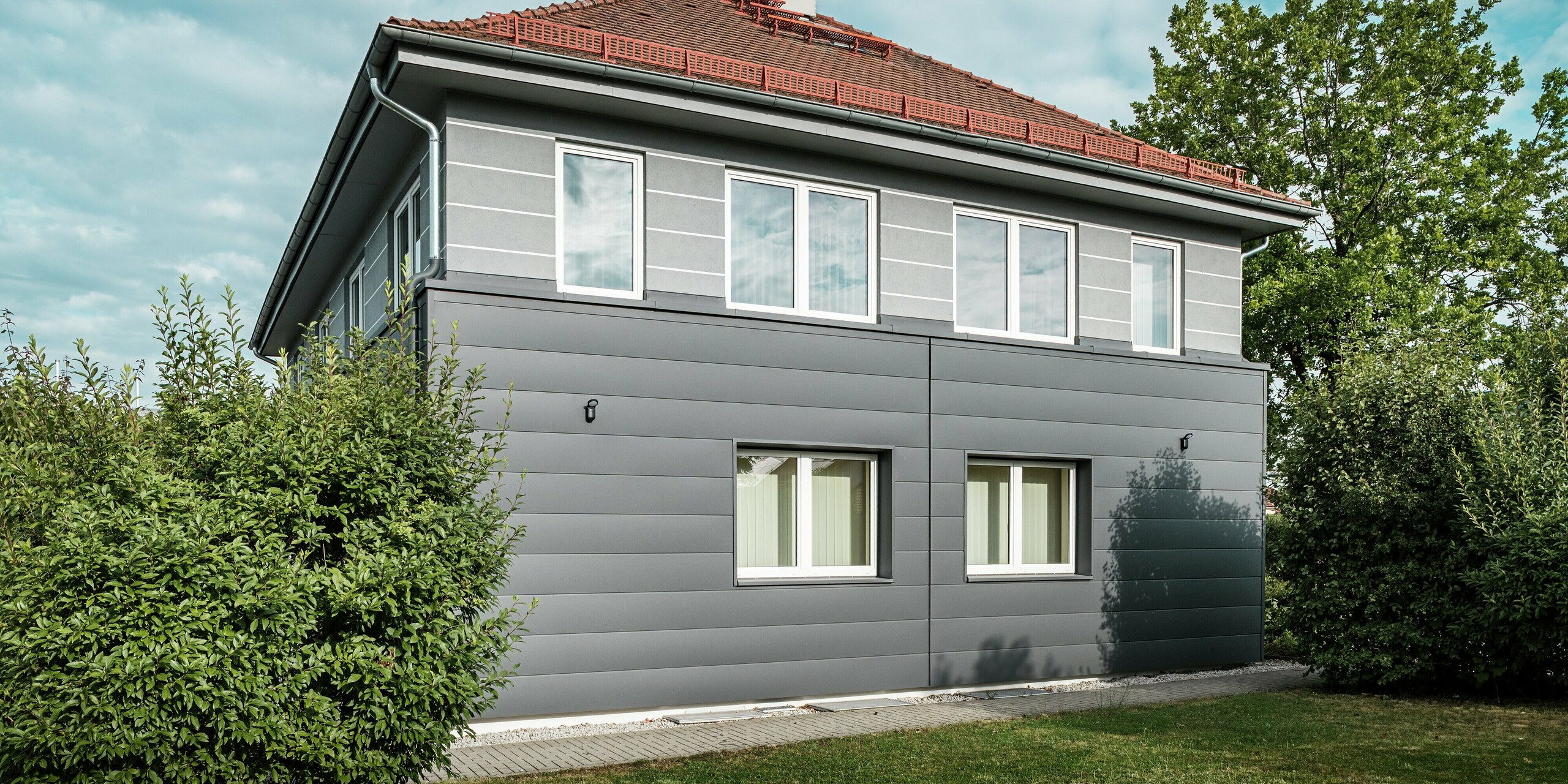 Das Bild zeigt die frisch sanierte Fassade der Kaminkehrerinnung in Bindlach, die mit den modernen PREFA Sidings in der eleganten Farbe P.10 Dunkelgrau ausgestattet wurde. Die klar strukturierten Aluminium-Paneele verleihen dem Gebäude eine zeitgemäße, stilvolle Optik und schützen es gleichzeitig effektiv vor Witterungseinflüssen. Besonders hervorzuheben ist die langlebige, pflegeleichte Oberfläche, die die Fassade stets in einwandfreiem Zustand hält. Die Verbindung von Funktionalität und modernem Design macht die PREFA Fassadenverkleidung zu einer idealen Wahl für anspruchsvolle Sanierungen.