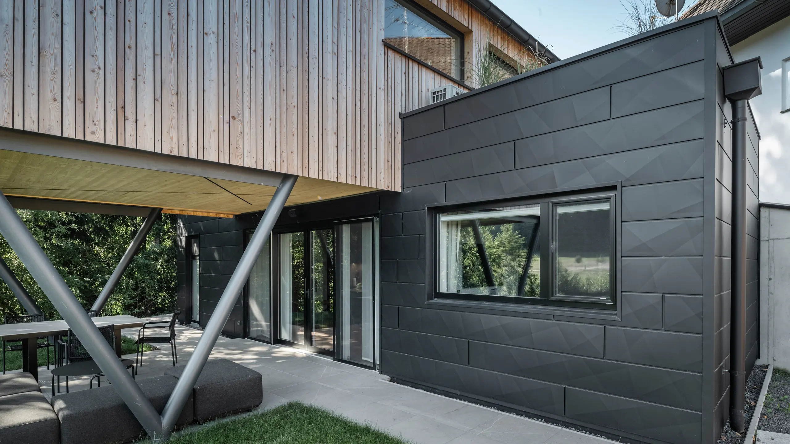 Arkitektonisk høydepunkt: «Woodstock Chalet 5» i St. Martin am Innkreis, Øvre Østerrike. Det moderne bryllupsstedet fremheves ved bruk av antrasittfargede PREFA Siding.X fasadepaneler. Aluminiumet danner en sterk kontrast til de lyse trepanelene og de metalliske støttestrukturene. De mørke vindusrammene passer harmonisk inn i fasadedesignet, noe som gir bygningen et elegant utseende. Dette bildet viser den estetiske balansen mellom naturlig tretekstur og den industrielle sjarmen til aluminiumsfasaden, som står for designet av moderne bygninger.
