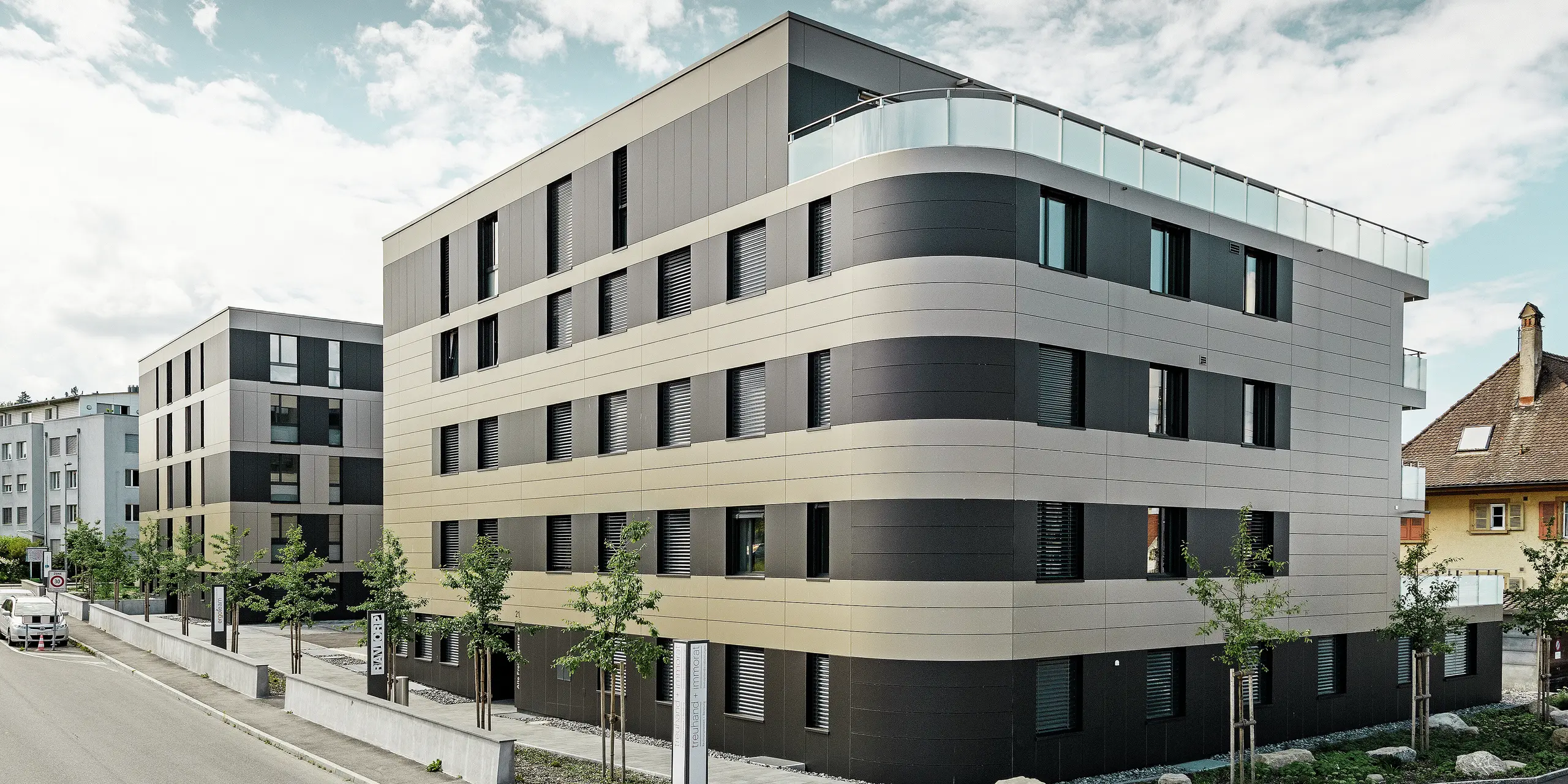 In der kubischen Wohnanlage von Murten (CH) sorgen die PREFA Aluminium Verbundplatten in Bronze und Schwarzgrau für eine optische Aufwertung und gleichzeitig für eine robuste und langlebige Fassade. Die Kombination dieser Farbtöne bietet ästhetische Akzente und schützt das Gebäude langfristig vor Wettereinflüssen. An der Gebäudeecke zur Straße hin ist eine Rundung zu erkennen, was das Rollbiegen der PREFABOND Aluminium Verbundplatten demonstrieren soll.