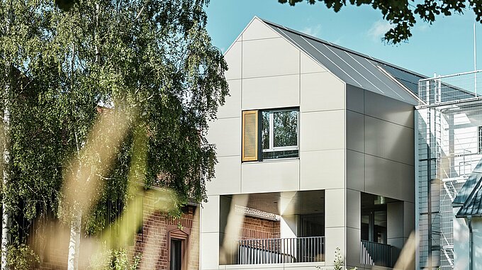 Das Bild zeigt das moderne Rathaus in Alzenau, dessen Fassade aus Aluminium-Verbundplatten von PREFA in einem eleganten Bronze-Ton besteht. Der spitze Dachbereich und die großen Fenster betonen den zeitgenössischen Charakter des Gebäudes. Im Vordergrund rahmen Bäume und Grünflächen das Gebäude ein, wobei die weiche Beleuchtung und der klare Himmel den modernen urbanen Raum unterstreichen.