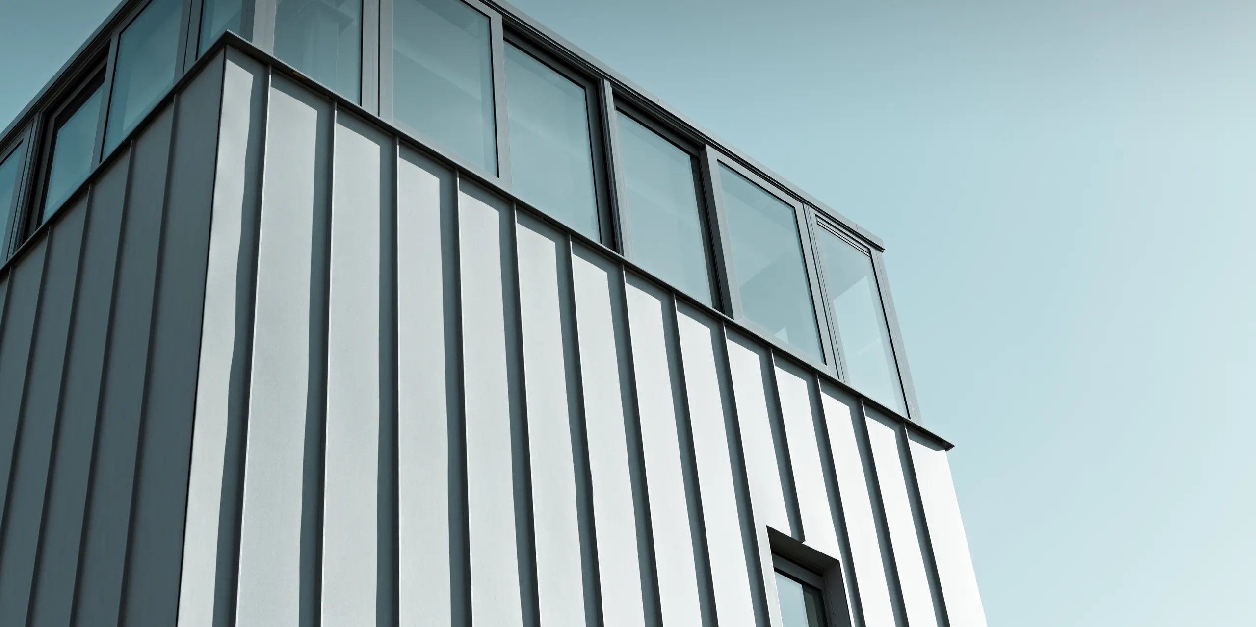Dynamische Detailaufnahme einer modernen PREFALZ Fassade in Patinagrau. Die klare Linienführung der vertikalen Stehfalzbahnen trifft auf großzügige Verglasung im Obergeschoss, die Licht und Transparenz schafft. Die langlebige PREFA Aluminiumfassade sorgt für dauerhaften Schutz und setzt architektonisch ein markantes Statement.