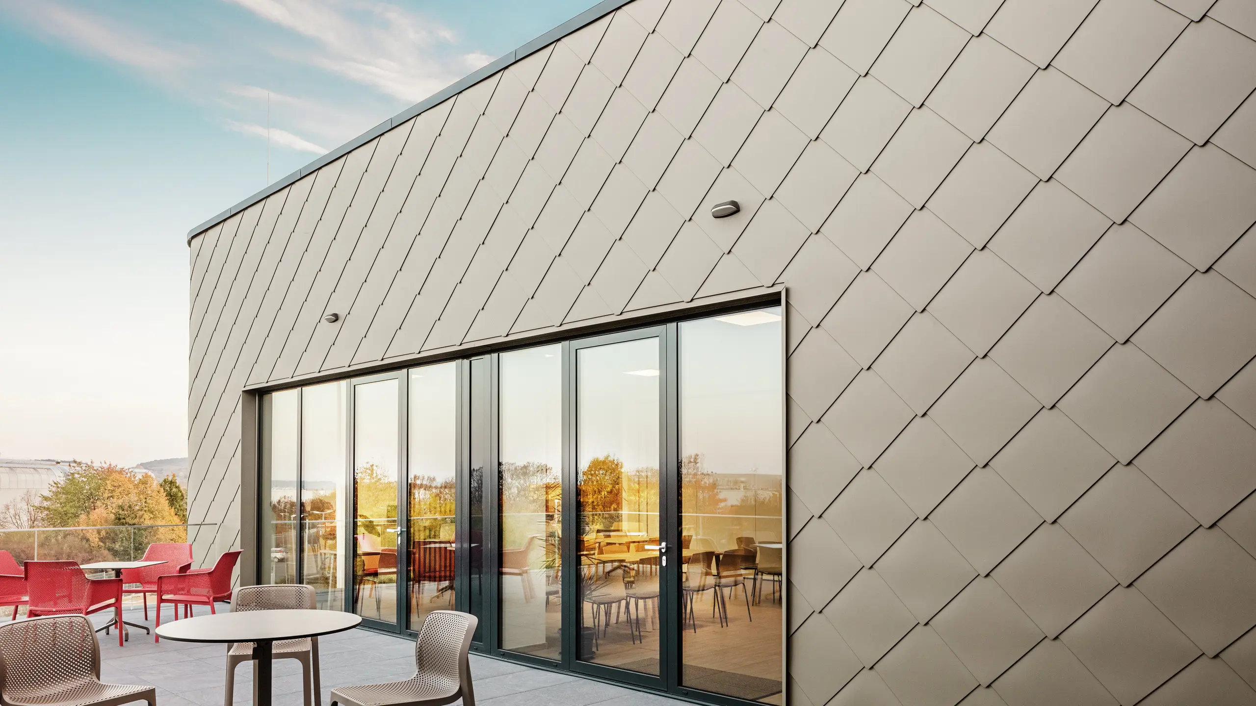 Die Fassade des neuen PREFA Standortes in Törökbálint besticht durch die eleganten PREFA Wandrauten 44 × 44 in P.10 Prefabronze. Die präzise verlegte Aluminiumfassade sorgt für ein modernes, dynamisches Erscheinungsbild und bietet gleichzeitig maximalen Schutz vor Witterungseinflüssen. Die großzügige Glasfront verbindet Innen- und Außenbereich und schafft auf der großzügigen Terrasse eine helle und einladende Atmosphäre.