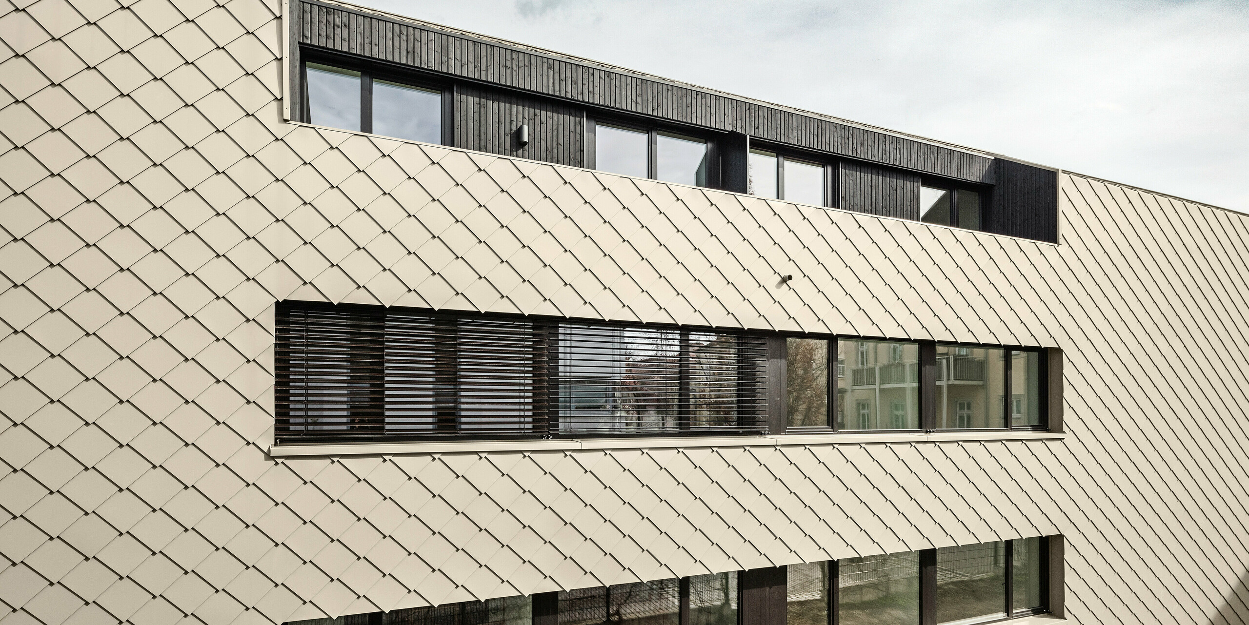 Rückansicht eines modernen Mehrfamilienhauses in Dresden-Plauen mit einer Fassade aus PREFA Wandrauten 29 × 29 in Bronze. Das Bild zeigt die gleichmäßig strukturierte Fassadenverkleidung mit großen, symmetrisch angeordneten Fenstern, die für viel Tageslicht in den Innenräumen sorgen. Die Kombination aus bronzefarbener Verkleidung und dunklen Fensterrahmen betont die durchdachte Bauweise des Gebäudes.