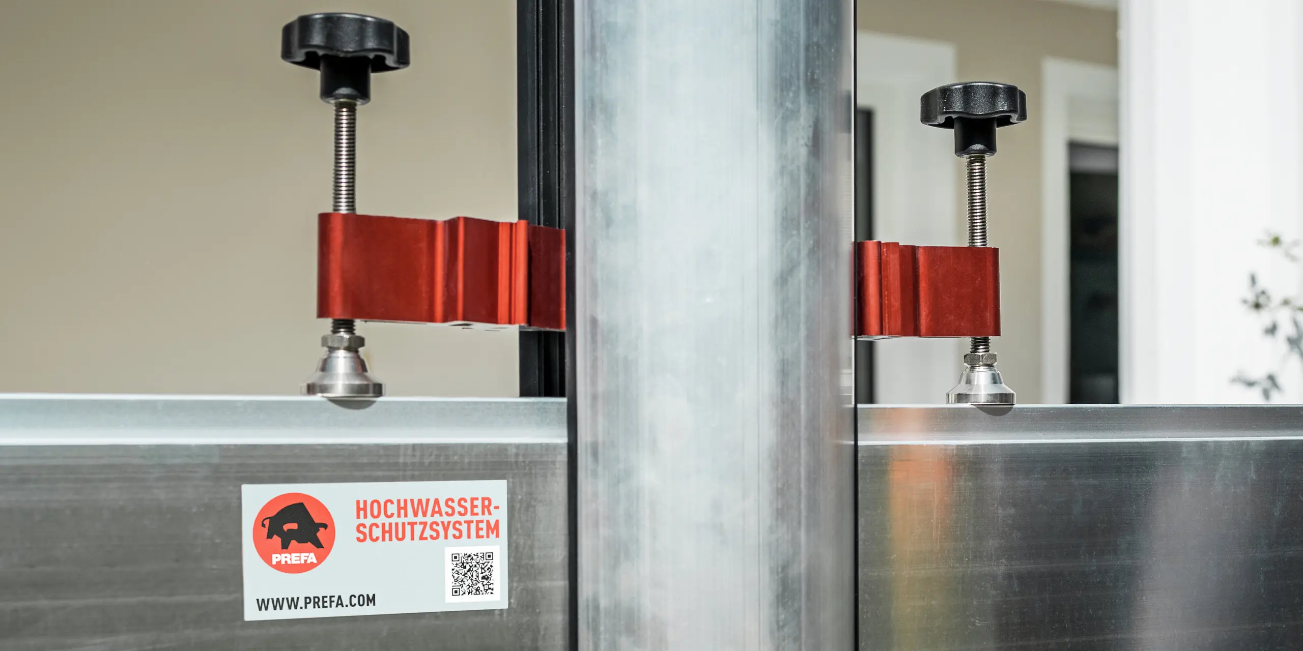 Nahaufnahme des PREFA Hochwasserschutzsystems mit Fokus auf die Spannvorrichtung zur Befestigung der Dammbalken. Gut zu erkennen sind die robusten Aluminiumprofile, die roten Spannelemente aus Metall und die schwarzen Handverschraubungen, die eine schnelle und sichere Montage ermöglichen. Das Bild zeigt die hochwertige Verarbeitung und Funktionalität des Systems - ein zentraler Baustein für den effektiven Hochwasserschutz von Gebäuden und Grundstücken.