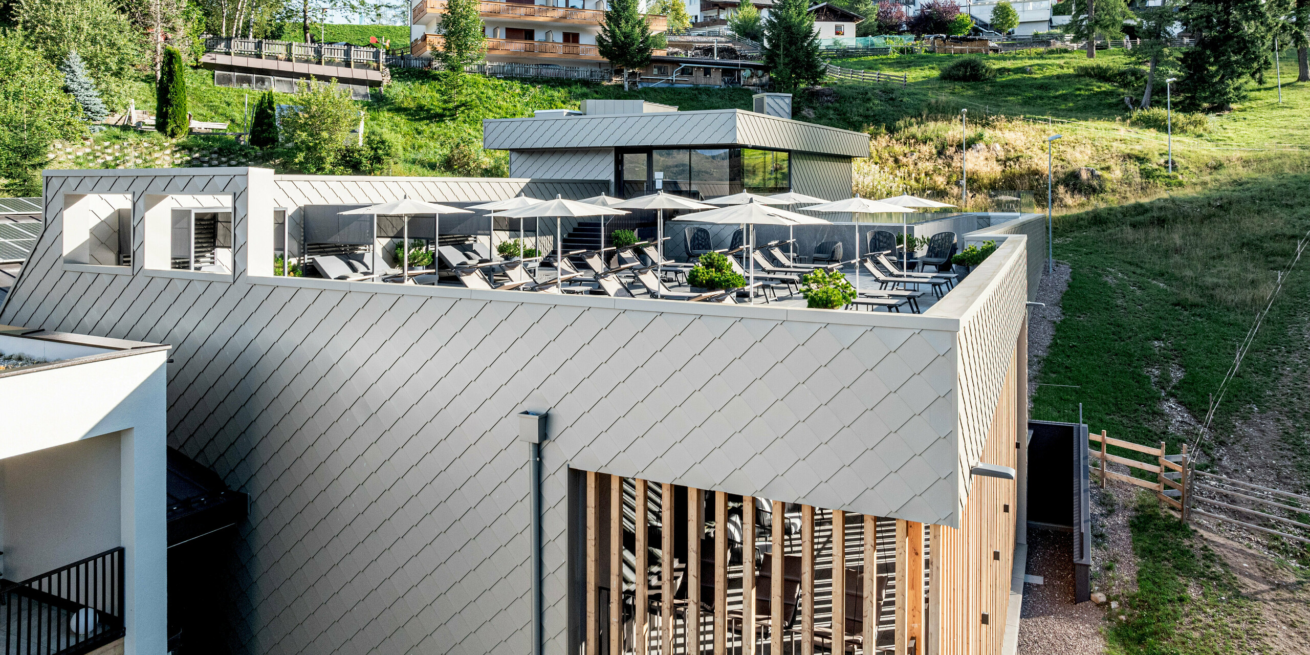 Erhöhte Terrasse des Sporthotels Obereggen in Südtirol mit Pool- und Loungebereich, Sonnenschirmen und freiem Blick auf die alpine Hanglandschaft und umliegende Chalets. Die klaren Baukörper werden von der PREFA Aluminiumfassade mit Wandraute 29 × 29 in der Farbe Bronze geprägt, die der Architektur eine präzise, lebendige Struktur verleiht. Der warme Bronzeton harmoniert mit Holz und Natur. Die langlebigen, witterungsbeständigen und wartungsarmen PREFA Aluminiumprodukte eignen sich ideal für moderne Hotelarchitektur im alpinen Raum.