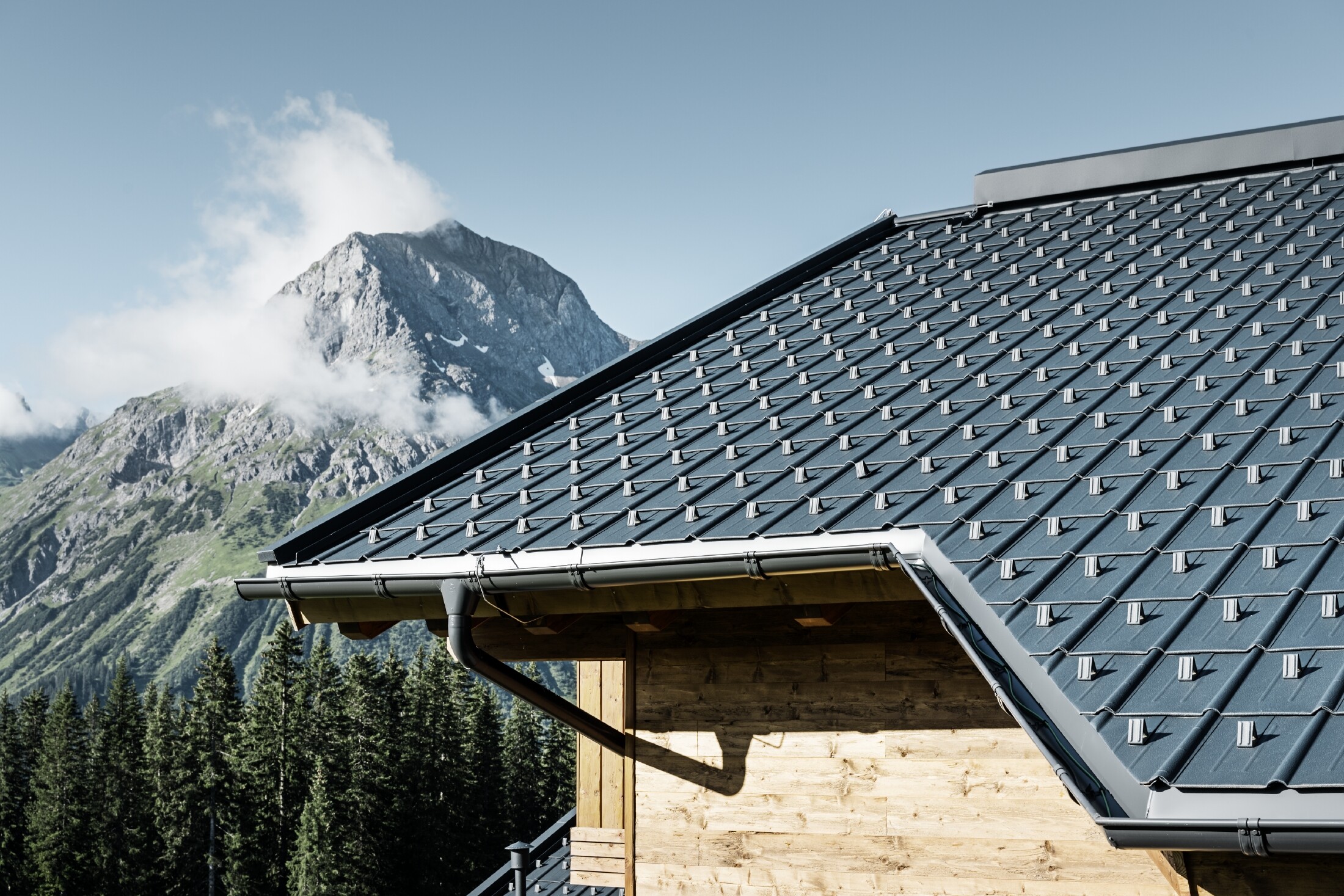 PREFA takpanel under alpine forhold - takpanel i antrasitt lagt på hytte i Lech am Arlberg med fjellene i bakgrunnen; PREFA takrenne i antrasitt; trefasade