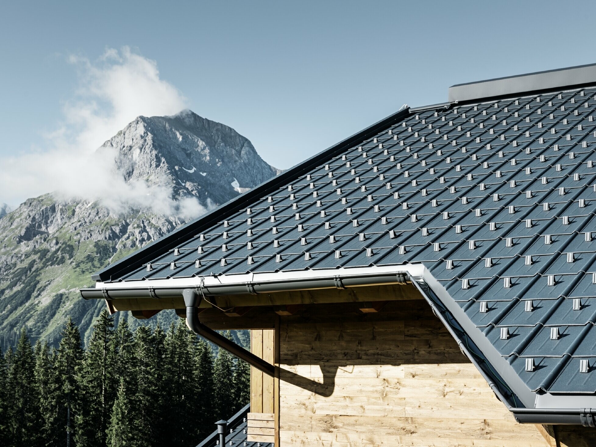 PREFA takpanel under alpine forhold - takpanel i antrasitt lagt på hytte i Lech am Arlberg med fjellene i bakgrunnen; PREFA takrenne i antrasitt; trefasade