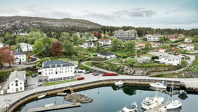 Flyfoto av havnen i Bekkjarvik, Norge, med Beckerwyc House i bakgrunnen. Hotellet skiller seg ut fra de omkringliggende tradisjonelle, hvite trehusene med sin futuristiske arkitektur og fasaden laget av PREFA-veggribber 44 × 44 i valnøttbrun. Det pittoreske landskapet, havnen med båtene og de grønne omgivelsene understreker hotellets unike beliggenhet i denne idylliske norske kystbyen på øya Selbjørn.