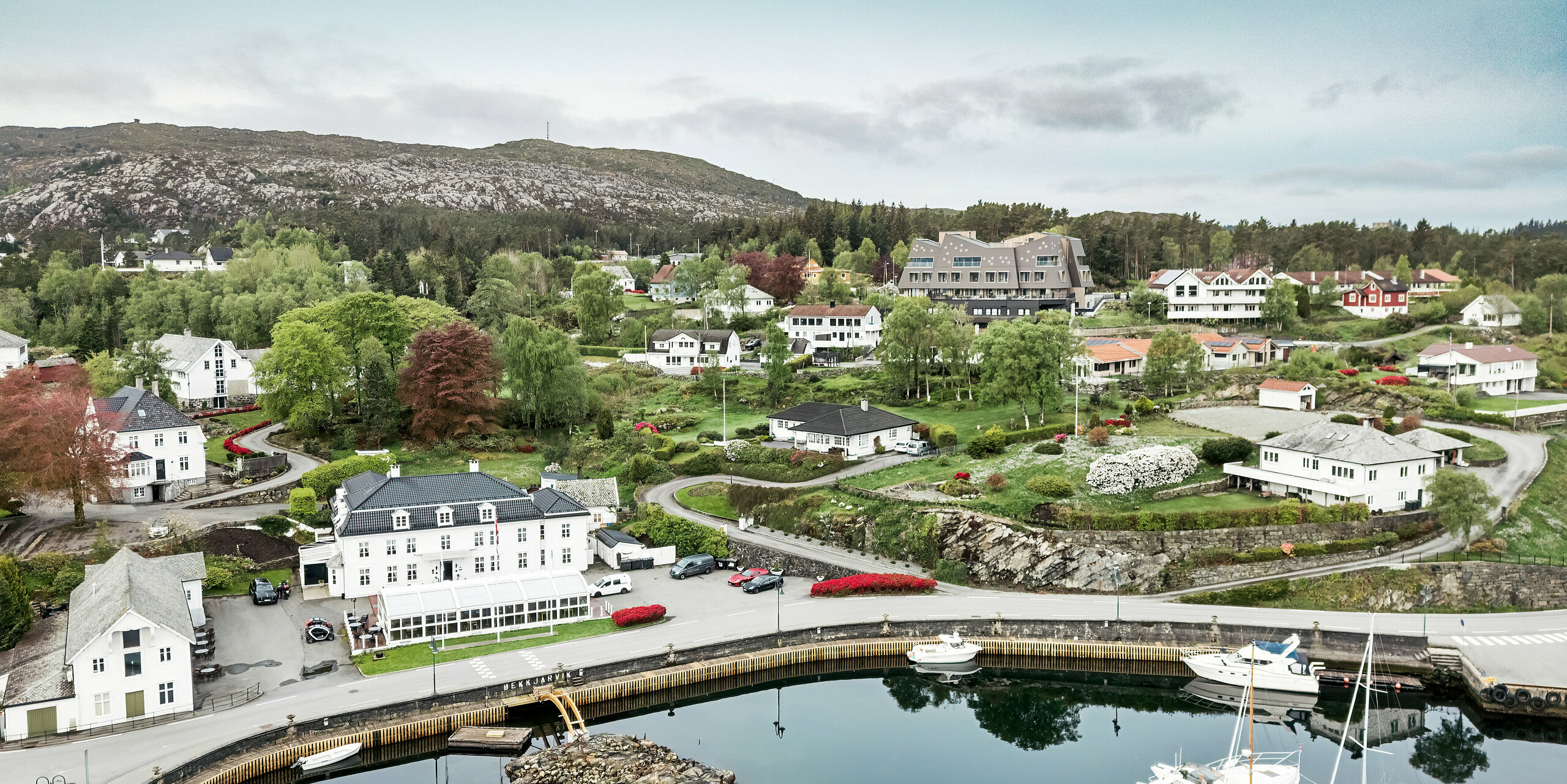 Flyfoto av havnen i Bekkjarvik, Norge, med Beckerwyc House i bakgrunnen. Hotellet skiller seg ut fra de omkringliggende tradisjonelle, hvite trehusene med sin futuristiske arkitektur og fasaden laget av PREFA-veggribber 44 × 44 i valnøttbrun. Det pittoreske landskapet, havnen med båtene og de grønne omgivelsene understreker hotellets unike beliggenhet i denne idylliske norske kystbyen på øya Selbjørn.