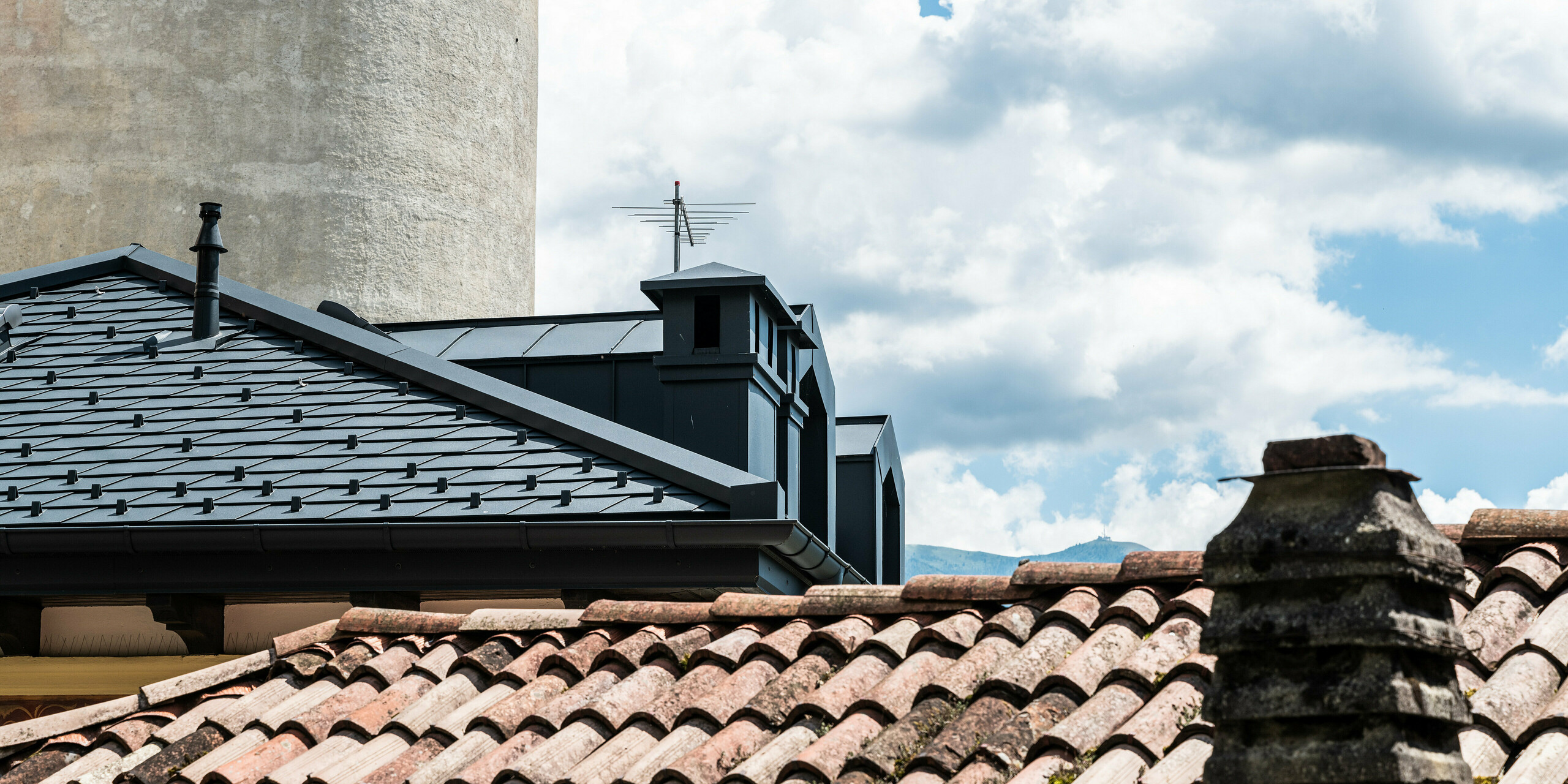 Kontrastreiche Ansicht eines renovierten Daches mit dunklen PREFA Dachschindeln in Anthrazit neben einem traditionellen Ziegeldach, hervorhebend, wie moderne Materialien die historische Architektur in Belluno, Italien, ergänzen. Das Bild fängt die Symbiose aus Alt und Neu ein, mit einem Fokus auf die Langlebigkeit und Ästhetik der PREFA Aluminiumprodukte im Einklang mit dem kulturellen Erbe der Umgebung.