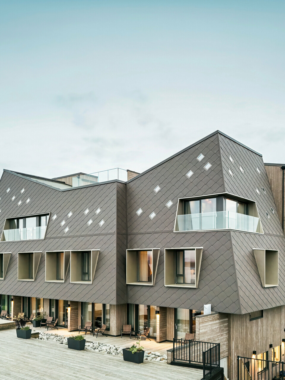 Utsikt over terrassen på hotellet Beckerwyc House i Bekkjarvik, Norge. Den moderne bygningen er utsmykket med en iøynefallende aluminiumsfasade laget av PREFA-veggribber 44 × 44 i valnøttbrun. Den futuristiske arkitekturen med skrånende takflater og utstikkende vinduselementer kompletteres av de romslige uteområdene. Fasadens naturlige farge passer harmonisk inn i det omkringliggende landskapet og understreker bygningens moderne design.